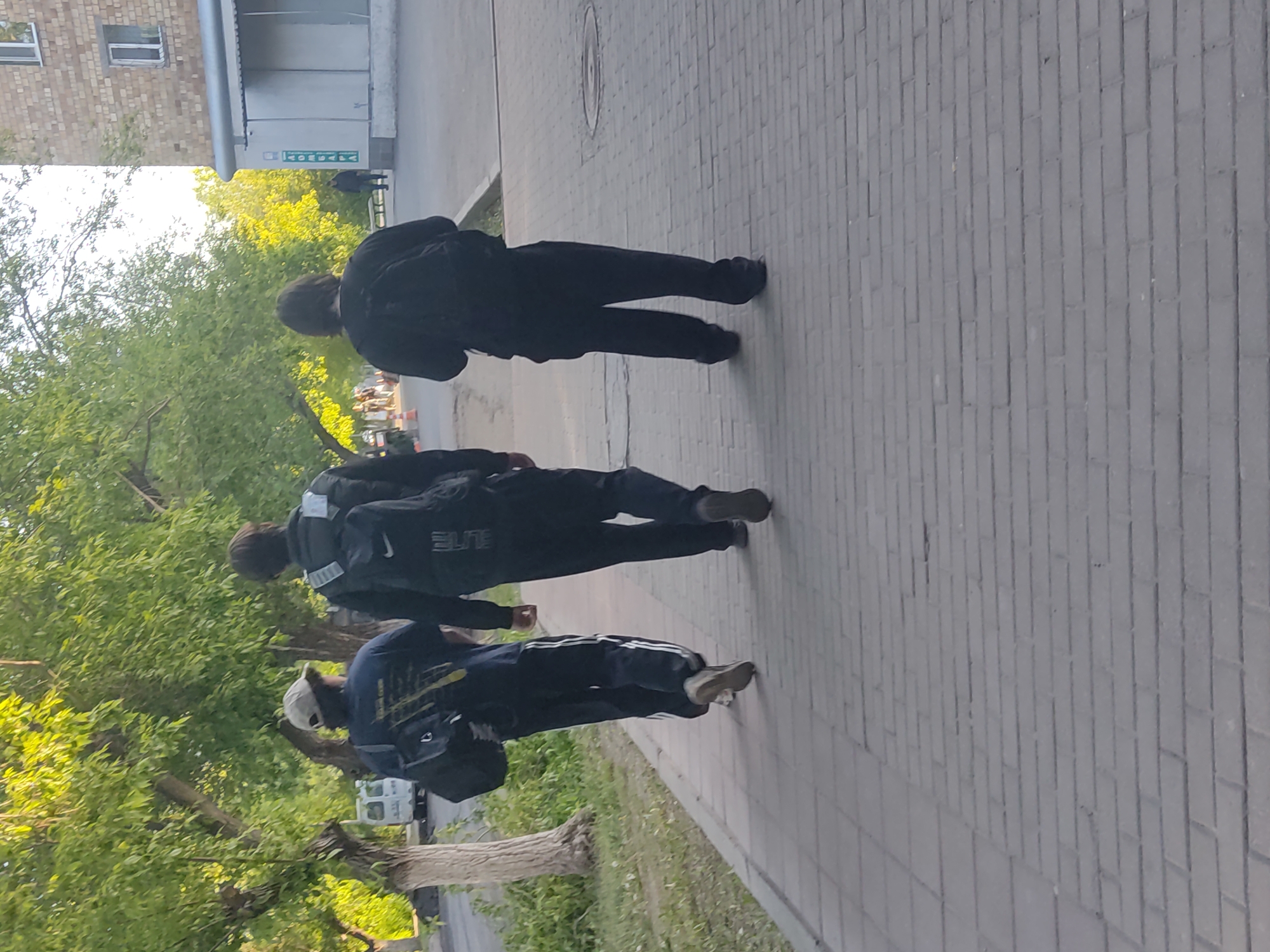 The image depicts three individuals walking along a brick sidewalk in an urban area. The person closest to the camera is wearing a navy blue tracksuit with white stripes on the pants, a light-colored cap, and a backpack. The second individual is wearing a dark-colored tracksuit with "ELITE" printed on the back. The third person is wearing all black clothing. Trees and greenery are visible to the left of the sidewalk. A building can be seen in the background.