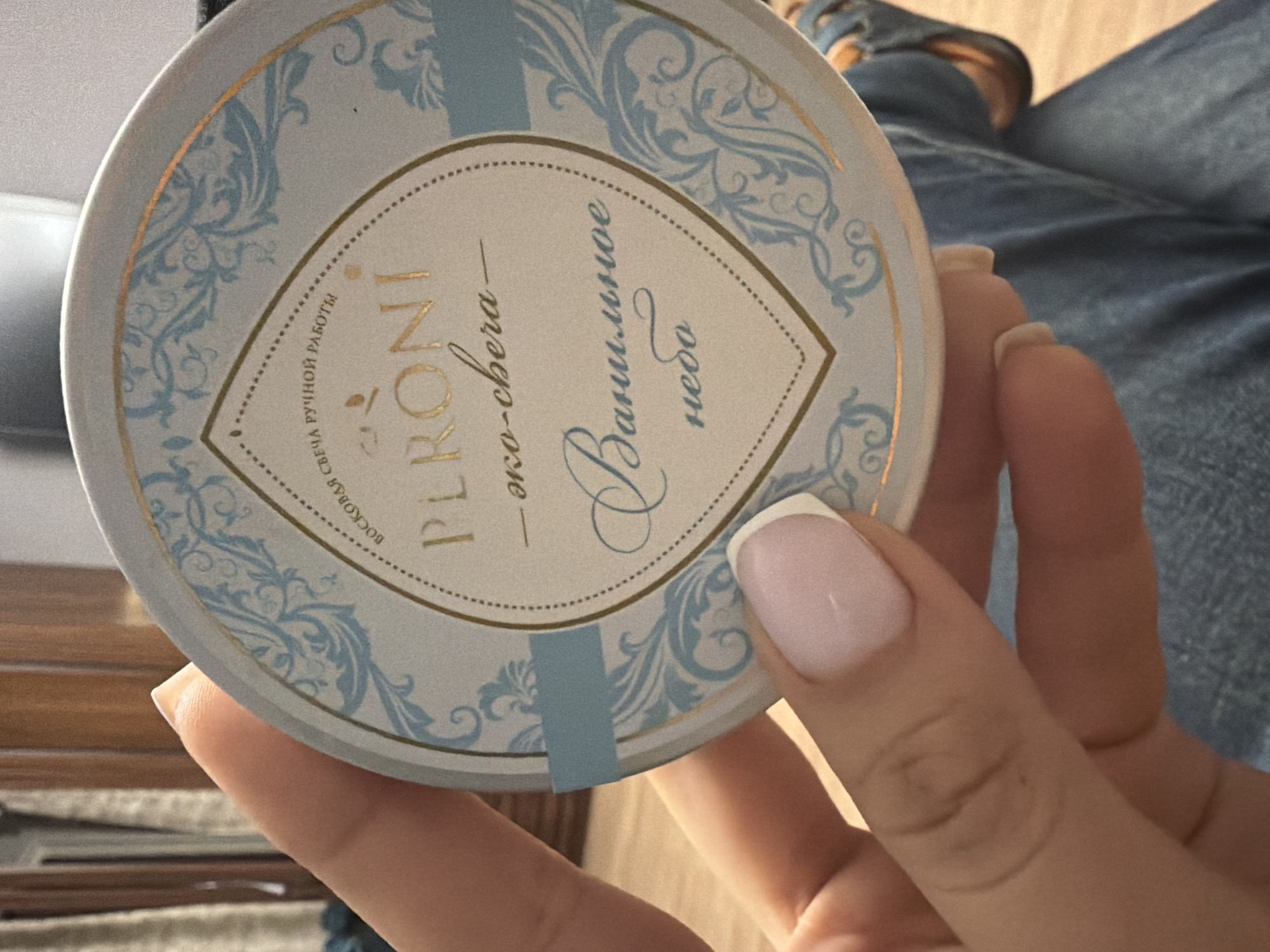 A person is holding a round, white tin with gold and light blue ornate designs. The lid of the tin displays text in Russian. The top line of text reads "ВОСКОВАЯ СВЕЧА РУЧНОЙ РАБОТЫ" which translates to "Handmade wax candle". Below that, "PLRÖNÍ" is written in large gold letters. Underneath "PLRÖNÍ", there is smaller text that reads "— еко-сhега —" and then "Ванильное небо" in a cursive font, translating to "Vanilla sky". The person's hand, with manicured nails, is holding the tin. The background shows a blurred interior setting with wooden furniture and denim fabric.