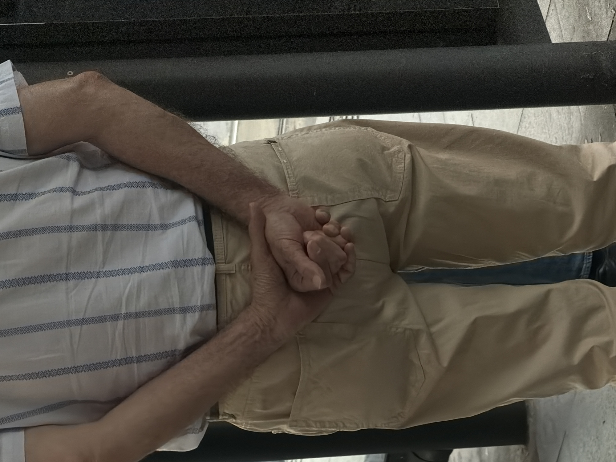 An individual is depicted from the rear, from the mid-torso to the upper legs, standing in an outdoor urban environment in Belgrade, Serbia. The person wears a light-colored, short-sleeved shirt with vertical blue patterned stripes, and tan-colored trousers. A dark belt is visible at the waistline. The shirt and trousers show signs of creasing and folds. The individual's hands are clasped behind their back, with fingers intertwined; visible veins and light body hair are present on the forearms and hands. To the right of the person, a dark cylindrical pole stands vertically, adjacent to a dark, vertical rectangular structure. The ground is paved with light-toned material. A second person's lower leg, wearing blue denim jeans and dark shoes, is partially visible behind and between the primary individual's legs.