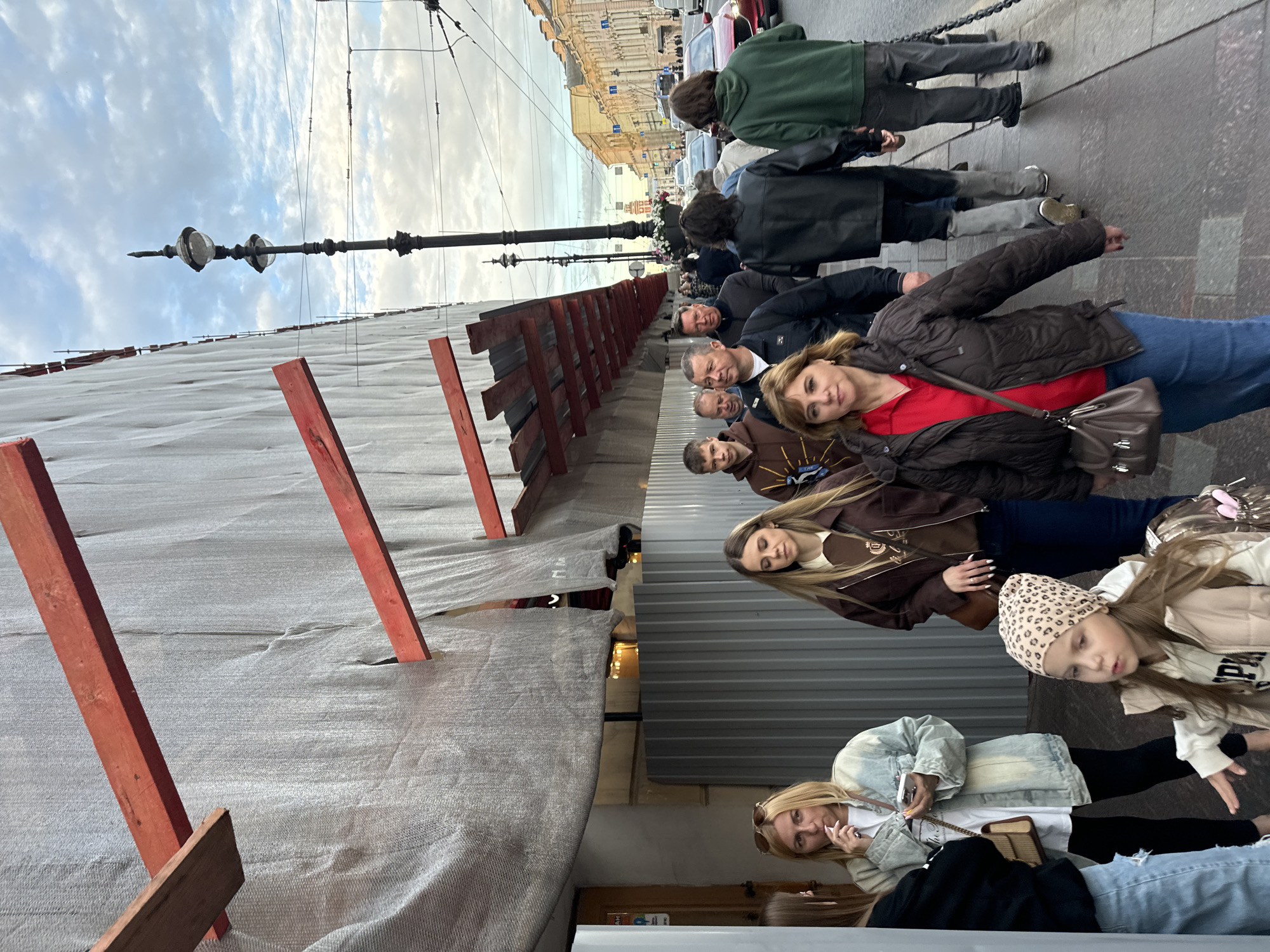 The image shows a group of people walking along a sidewalk in Saint Petersburg, Russia. The sidewalk is wet, suggesting it may have recently rained. The buildings on the right side of the image appear to be in a historical district, with ornate architecture. The building on the left is under construction or renovation, covered by a grey tarp and supported by scaffolding. The sky is cloudy, typical of the city's climate. The people are dressed in casual clothing, indicating it is likely daytime. Some individuals are looking at their phones, while others are engaged in conversation. The overall scene depicts a normal day in an urban environment.