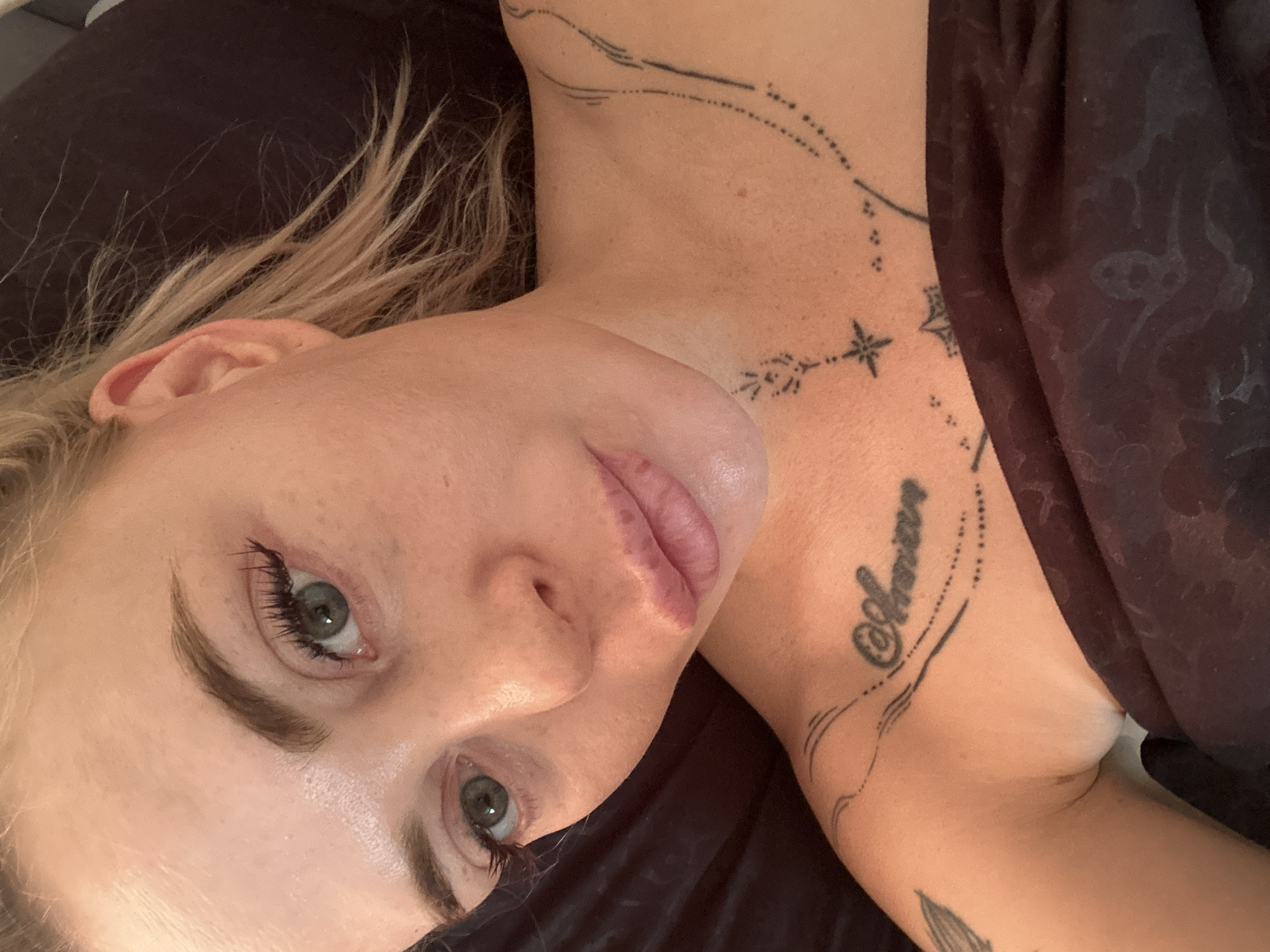 A close-up, overhead shot shows a fair-skinned person with blonde hair pulled back, lying down, looking towards the camera. The person's face is turned slightly to the left. They have blue-green eyes, light eyebrows, and visible pores and freckles on their skin. Their lips are slightly parted, and they are wearing subtle makeup. Tattoos are visible on the person's left shoulder and upper chest, including a script tattoo that reads "Amour" and a star design. The person is covered by a dark, patterned fabric, likely a blanket or bedding. The background is dark and out of focus. There are no discernible environmental details to suggest a specific city or location.