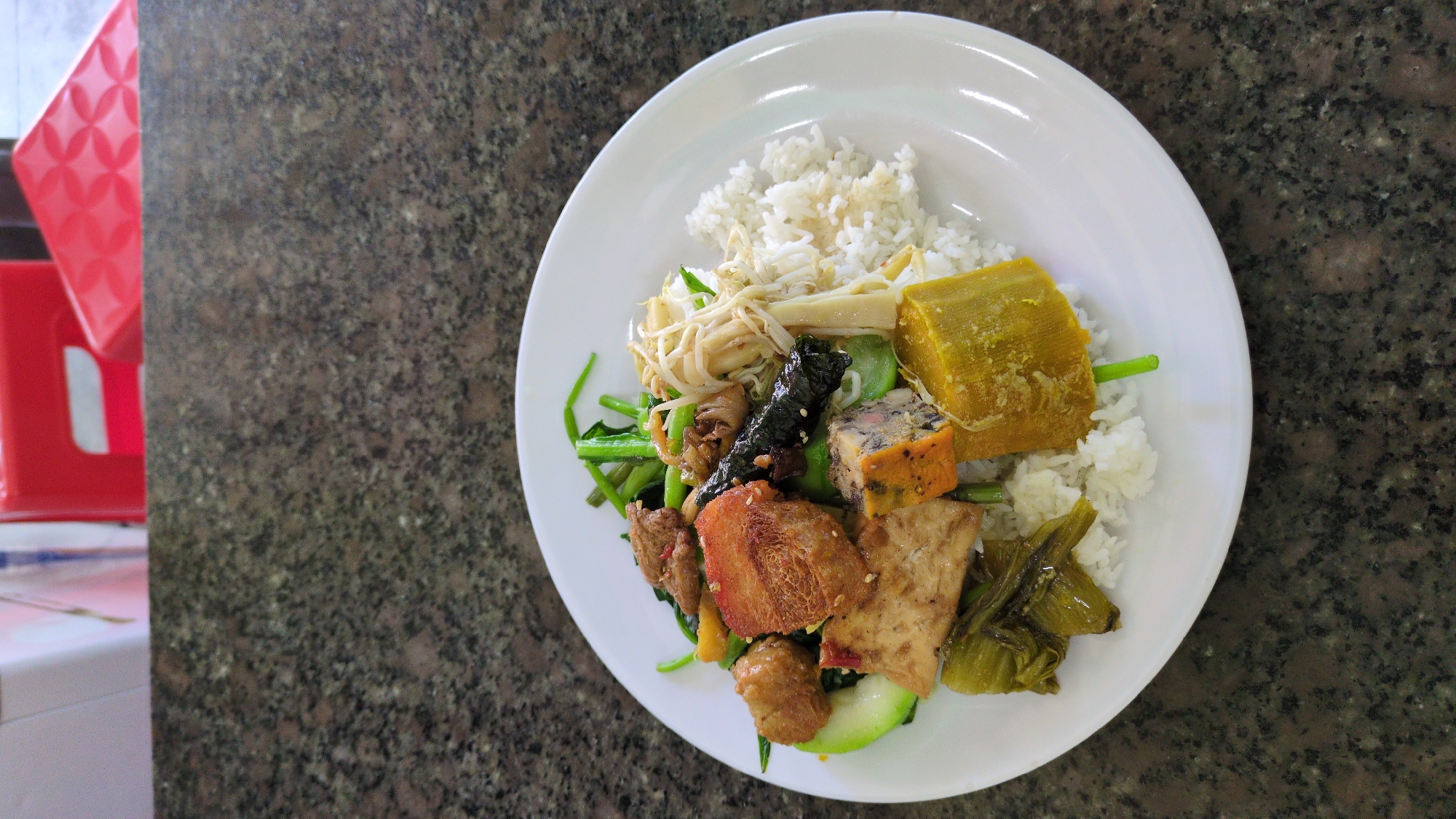 A plate of food sits on a speckled brown surface, possibly a table or countertop. The food appears to be a Southeast Asian meal, consisting of white rice topped with an assortment of items including a large cube of yellow squash, various meats (some fried, some glazed), tofu, green vegetables (likely water spinach), bean sprouts, bamboo shoots, seaweed, and a type of sliced egg loaf. A red, plastic chair is partially visible in the background. The location is likely a restaurant in South Quy Nhon Ward, Vietnam, based on the food type.