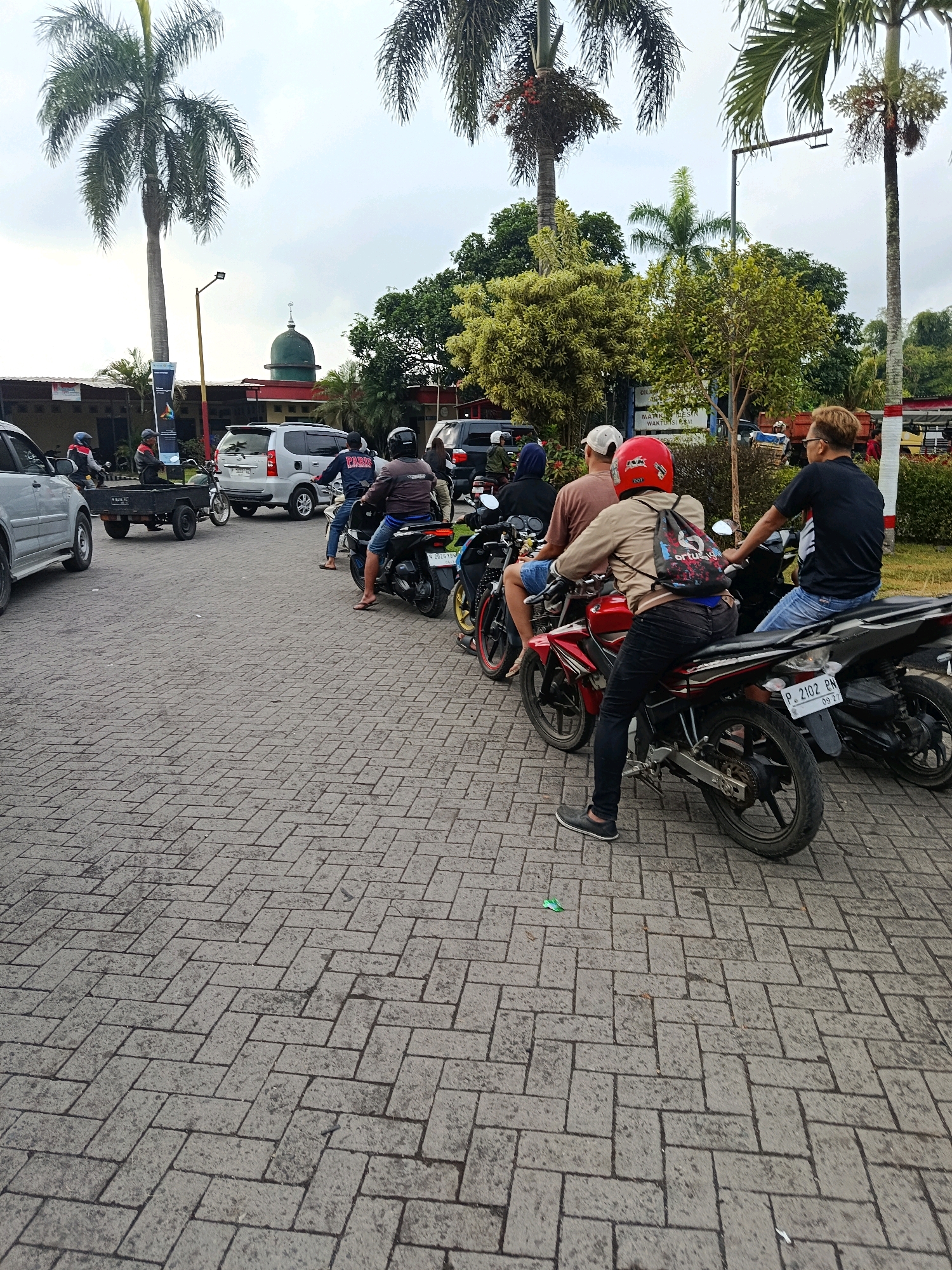 The image depicts a street scene in Tempeh Lor, Indonesia, where several people are seen on motorcycles and in cars, suggesting a moderate level of traffic or a gathering. In the foreground, the ground is paved with interlocking gray bricks. Two motorcycles are prominently featured, one red and one black, with riders seated on them. A red helmet with a black backpack is visible on the rider of the red motorcycle. A silver car is parked on the left side of the frame. In the background, a white van and a black car are present, along with a small trailer being pulled by a motorcycle. Further in the background, palm trees and other foliage are visible, as well as a building with a green dome, likely a mosque, and a street lamp. The sky is overcast.
