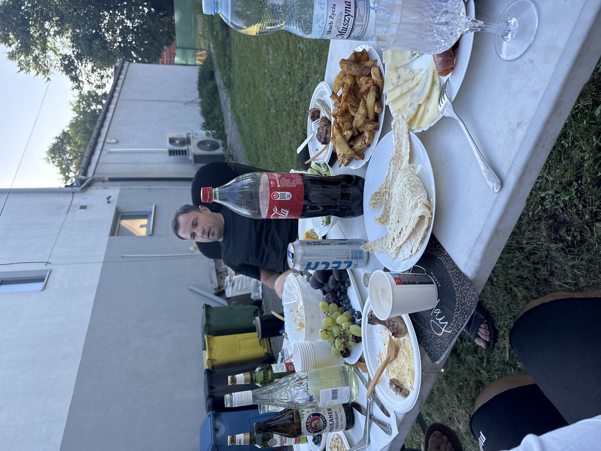 The image depicts an outdoor gathering at dusk, possibly in a backyard in Katowice, Poland. A group of people, one of whom is visible in the center of the frame, are seated around a white plastic table laden with food and drinks. The food includes plates of sausages, french fries, grapes, blueberries, cheese, and flatbread. Beverages on the table consist of bottles of beer, a large bottle of Coca-Cola, and a bottle of water. In the background, a building with grey and white siding is visible, along with several large trash bins and trees. The overall atmosphere suggests a casual meal or party.