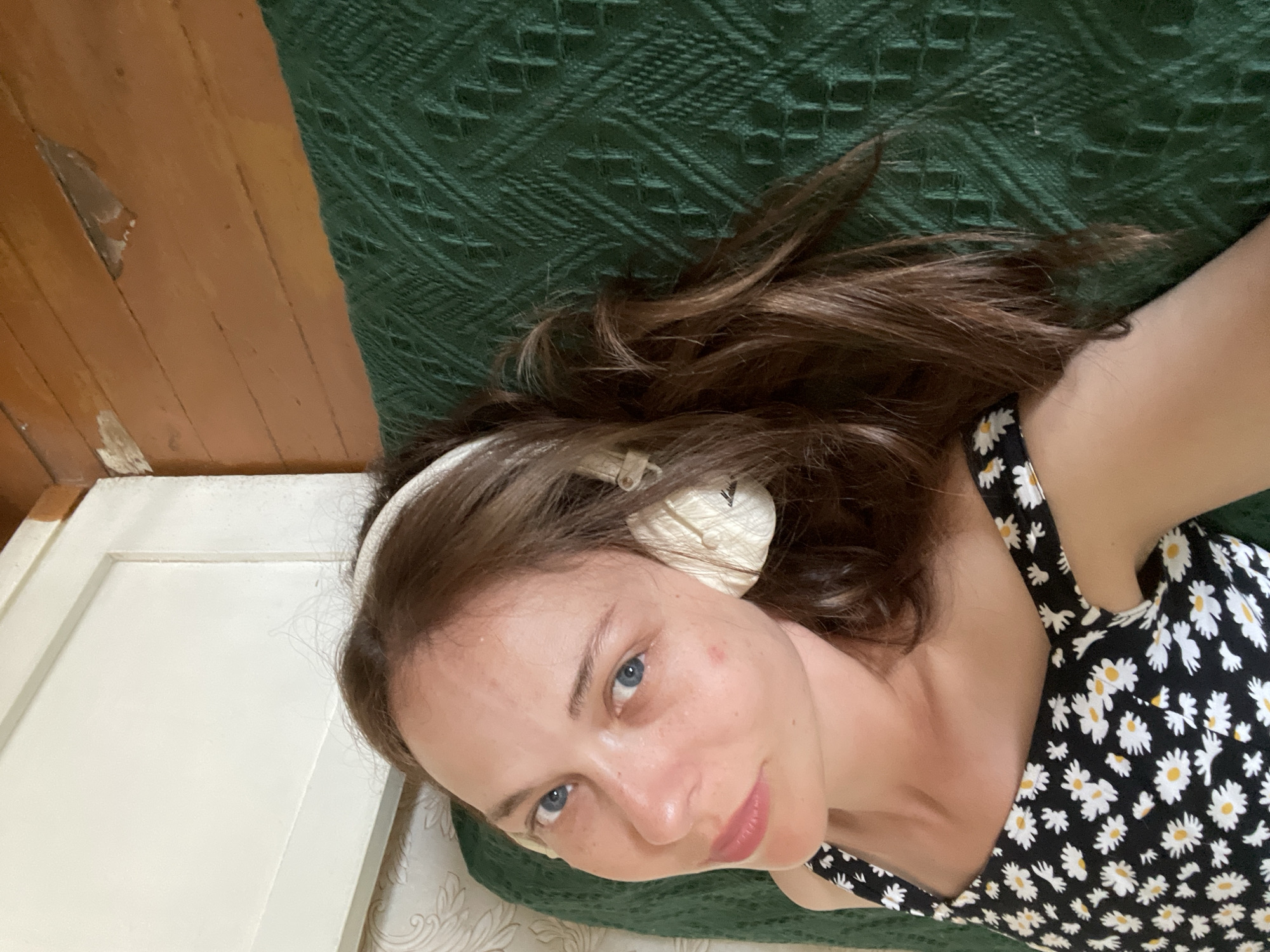 A young woman with blue eyes and brown hair is lying on her back on a green textured blanket. She is wearing a black dress with white daisies. She has on white headphones over her ears and her hair is spread out around her head. The background shows a wooden floor and a white door. The image is oriented so that the woman's head is at the top.