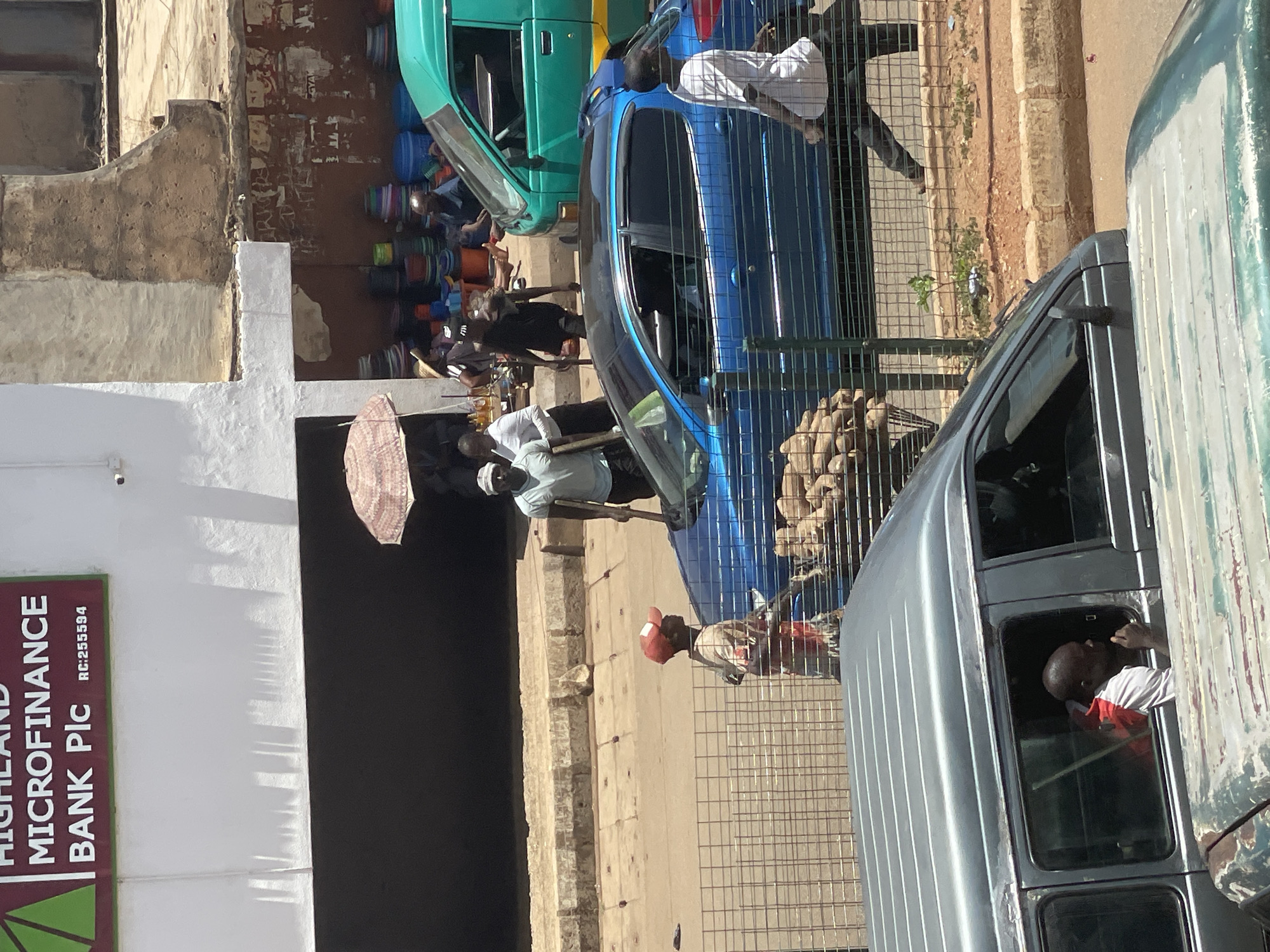 The image captures a street scene in Bukuru, Nigeria, adjacent to a building housing "Highland Microfinance Bank Plc" with RC:255594 displayed. People are walking on the sidewalk. Several vehicles are parked or moving along the road. There are several people outside. One person is holding a pink umbrella. Merchandise, including colorful buckets, are visible. A man is looking out the window of the right-most vehicle. A wire fence is on the right.