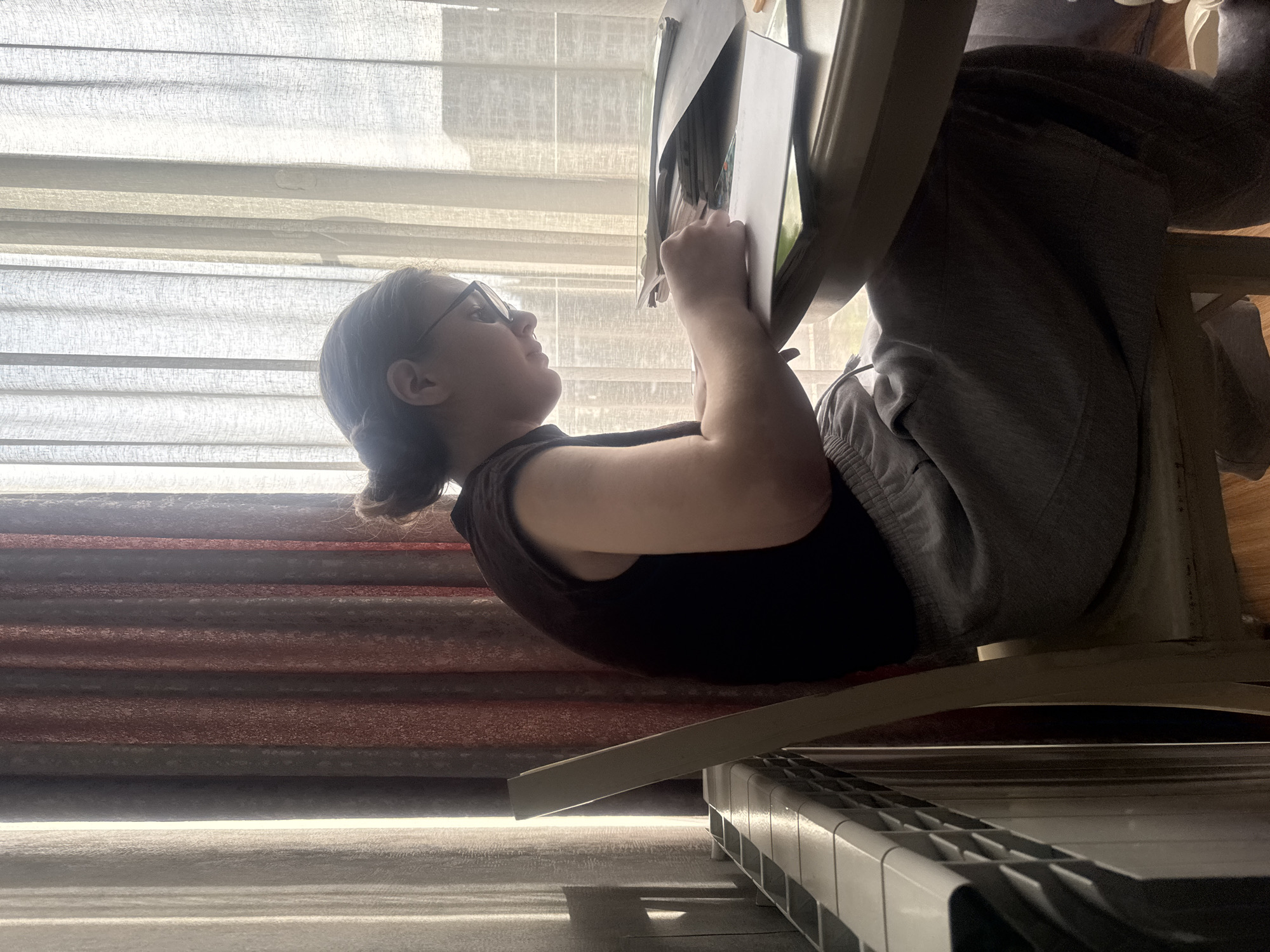 The image, correctly oriented, displays an indoor scene featuring a young female engaged in an activity. She is seated in what appears to be a chair, facing towards a large window covered by translucent white horizontal blinds. Bright daylight streams through the window, backlighting the subject and the room.
The female subject has dark hair tied back and is wearing glasses. She is dressed in a dark, sleeveless top and grey sweatpants. Her arms are raised, and she holds open what appears to be a book or sheet music, which is resting on a stand or table in front of her. Her gaze is directed upwards at the material, suggesting she is reading, studying, or possibly practicing.
In the bottom right corner of the frame, a segment of a digital piano or keyboard is visible, showing its white and black keys. The floor beneath the subject and extending towards the keyboard features horizontal stripes in shades of grey and reddish-brown, possibly a rug or part of the flooring.
The bright illumination from the window indicates it is daytime. The overall impression is one of quiet concentration. No visible text is legible in the image.