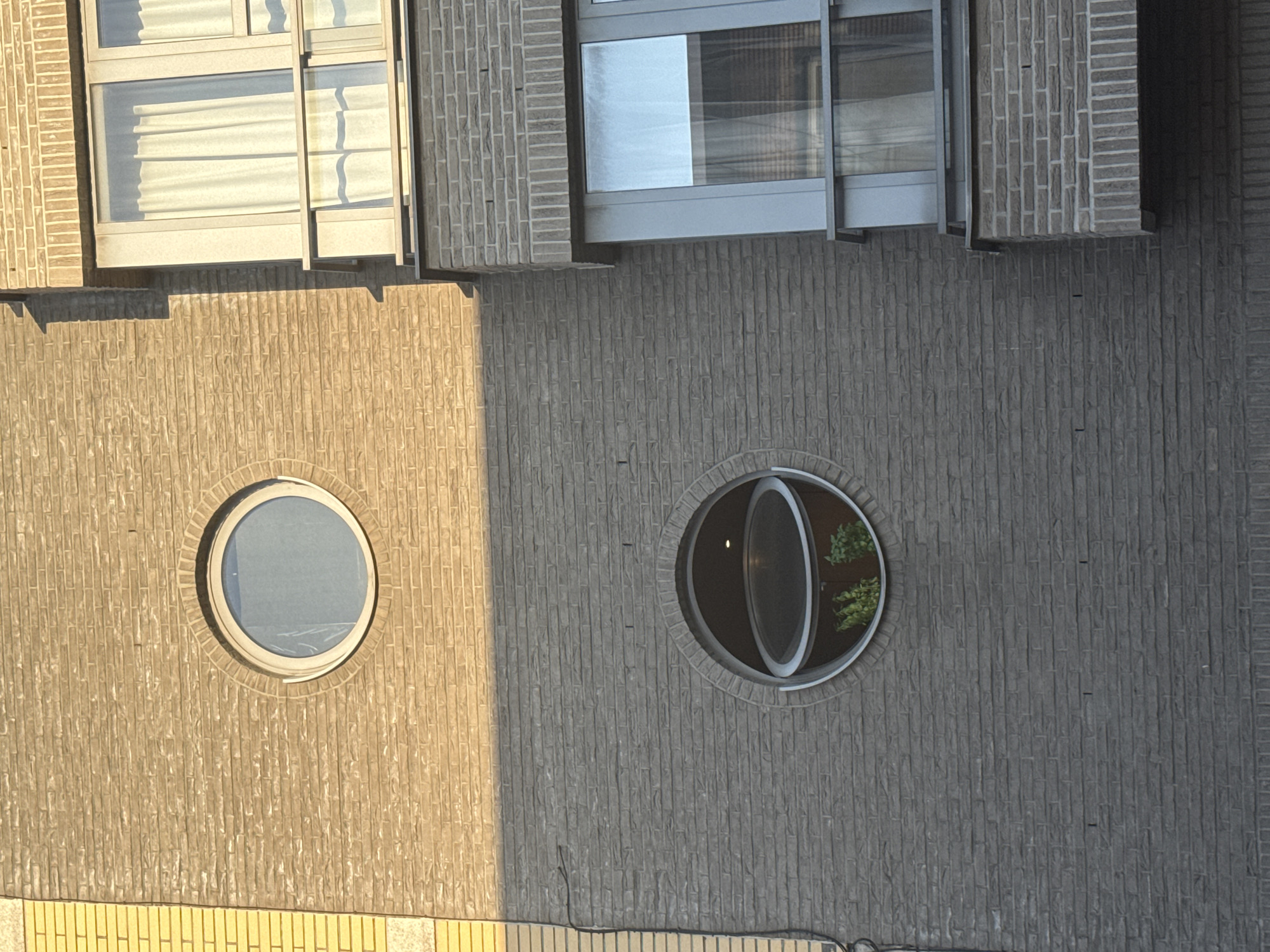 The image shows a close-up, exterior view of a modern brick building with two distinct styles of brickwork. The upper portion of the building features light brown, textured brick, while the lower section is constructed with darker gray, rectangular bricks. Two circular windows are visible: one near the top of the light brown section, which is a closed, circular pane of glass, and another in the dark gray section, which is a circular opening with a hinged section of glass propped open, revealing greenery inside. To the right of the circular windows, a modern section of the building with large, rectangular windows and a balcony is visible. The overall impression is one of contemporary architecture with unique window designs, located in Strombeek-Bever, Belgium. There are no people or animals present in the image. The lighting suggests it is daytime, with sunlight casting shadows on the building's facade.