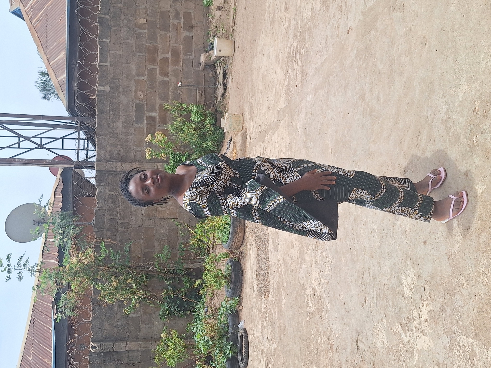 A full-body, portrait-oriented shot captures a woman standing outdoors under bright daylight in Sabon Tasha, Nigeria. She is positioned slightly to the left of the frame, looking directly at the camera with a neutral expression. She wears a long-sleeved, ankle-length dress or gown featuring a green, black, and white geometric pattern, and light pink flip-flops. Her dark hair is styled in braids.
The ground beneath her is a light-colored, dusty or sandy surface. Behind her, a low-growing plant with light green leaves and small white flowers is visible, with other plants seemingly growing in several repurposed tires acting as planters. A brick wall, likely part of a building or a boundary, stretches horizontally across the mid-ground. Along the top of this wall, a line of razor wire is present.
In the background, a corrugated metal roof is visible on the left, along with a grey metal structure, possibly part of an antenna or a support frame. A white satellite dish is mounted high up, near the roofline on the left. On the right side, two light-colored buckets are placed near the base of the brick wall. The sky is clear, indicating a sunny day. There is no visible text in the image.