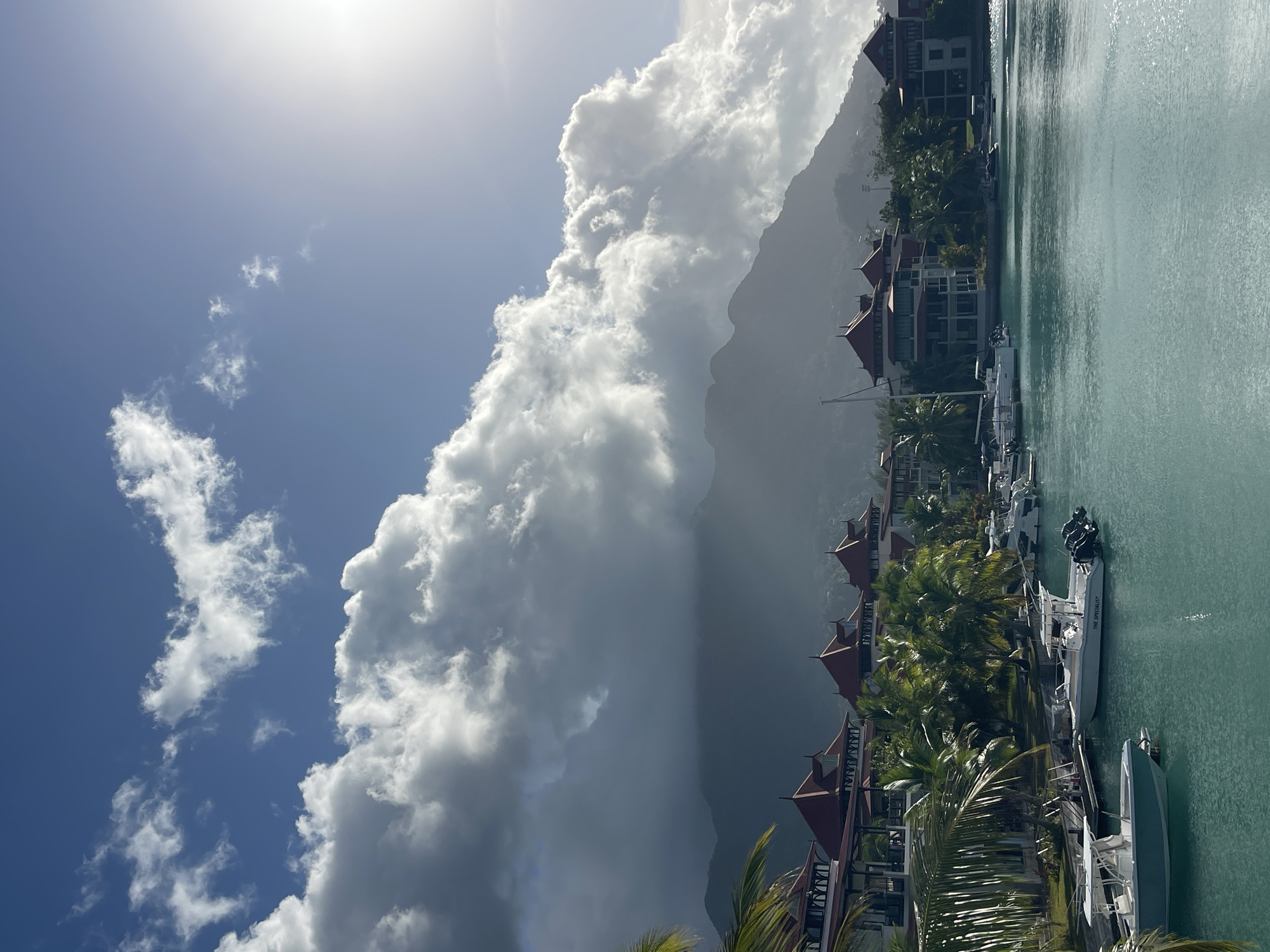 The image shows a picturesque waterfront scene in Victoria, Seychelles, under a dramatic sky. In the foreground, calm, turquoise water is dotted with several white motorboats docked along a pier. The boats are detailed, with visible engines and seating areas. Beyond the water, a line of modern buildings with red-tiled roofs and balconies stretches along the shore, interspersed with lush palm trees. These buildings appear to be part of a resort or residential complex. In the background, a large, mist-shrouded mountain range rises, its peaks partially obscured by heavy, white and gray clouds. The sun is visible in the upper right corner, casting a bright glare and highlighting the edges of the clouds. The overall impression is one of a serene tropical location with dynamic weather conditions.