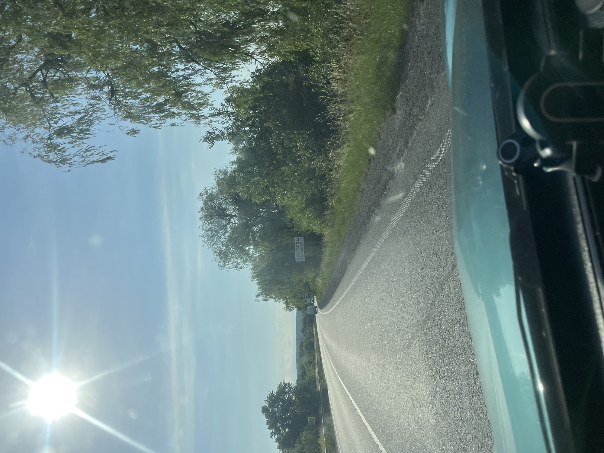 The image is a view from the driver's seat of a car on a sunny day, looking towards a road that curves to the left. A sign is visible on the right side of the road with Cyrillic text that reads "УЖГОРОД UZHHOROD". This indicates the car is approaching or is in the vicinity of Uzhhorod. The road has two lanes, with white lines marking the lanes. The surrounding environment is lush and green, with trees lining the road and grass growing along the roadside. The sky is blue with some wispy clouds and the sun is bright, causing a lens flare effect in the upper left corner of the image. There are a few vehicles visible further down the road. The dashboard of the car, colored in a teal hue, is visible in the foreground.