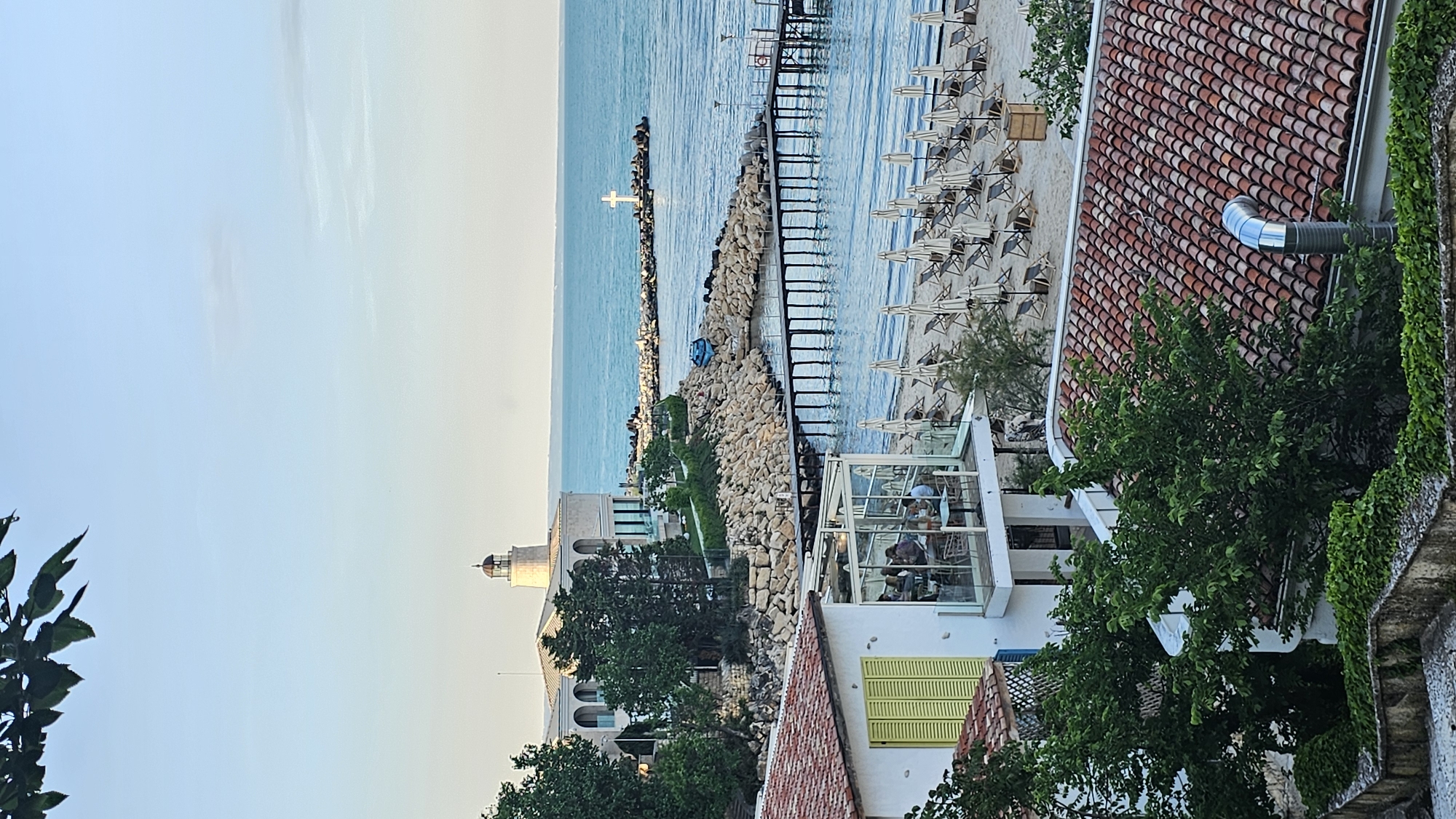 The image depicts a coastal scene in Varna, Bulgaria. The foreground is dominated by rooftops with red terracotta tiles, some partially obscured by lush green trees and climbing ivy. A white building with yellow-green shutters and a glass-enclosed dining area is visible. Through the glass, several individuals are seated at tables, suggesting a restaurant setting. Beyond the rooftops, a narrow sandy beach stretches along the coastline, lined with numerous white sun umbrellas and lounge chairs. A stone breakwater extends into the turquoise sea, culminating in a white cross. A small structure resembling a lighthouse is also visible near the beach. The sky is a gradient of light blue, fading into a lighter hue near the horizon.