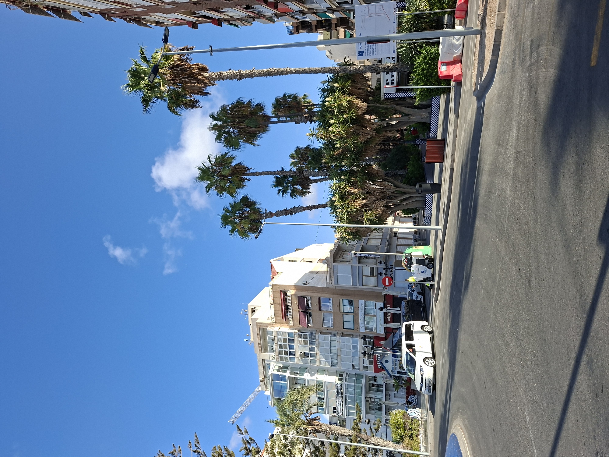 The image depicts a street scene in Torrevieja, Spain, under a clear blue sky with scattered white clouds. A white van is driving on the asphalt road, which curves to the left. To the right of the van, a street cleaning machine is visible. In the background, several multi-story buildings stand, one of which is adorned with balconies and awnings. Palm trees and other lush greenery are scattered throughout the scene, contributing to a tropical or Mediterranean atmosphere. The buildings are a mix of modern and traditional architecture. Streetlights and utility poles are also present. The overall impression is that of a sunny day in a coastal city.