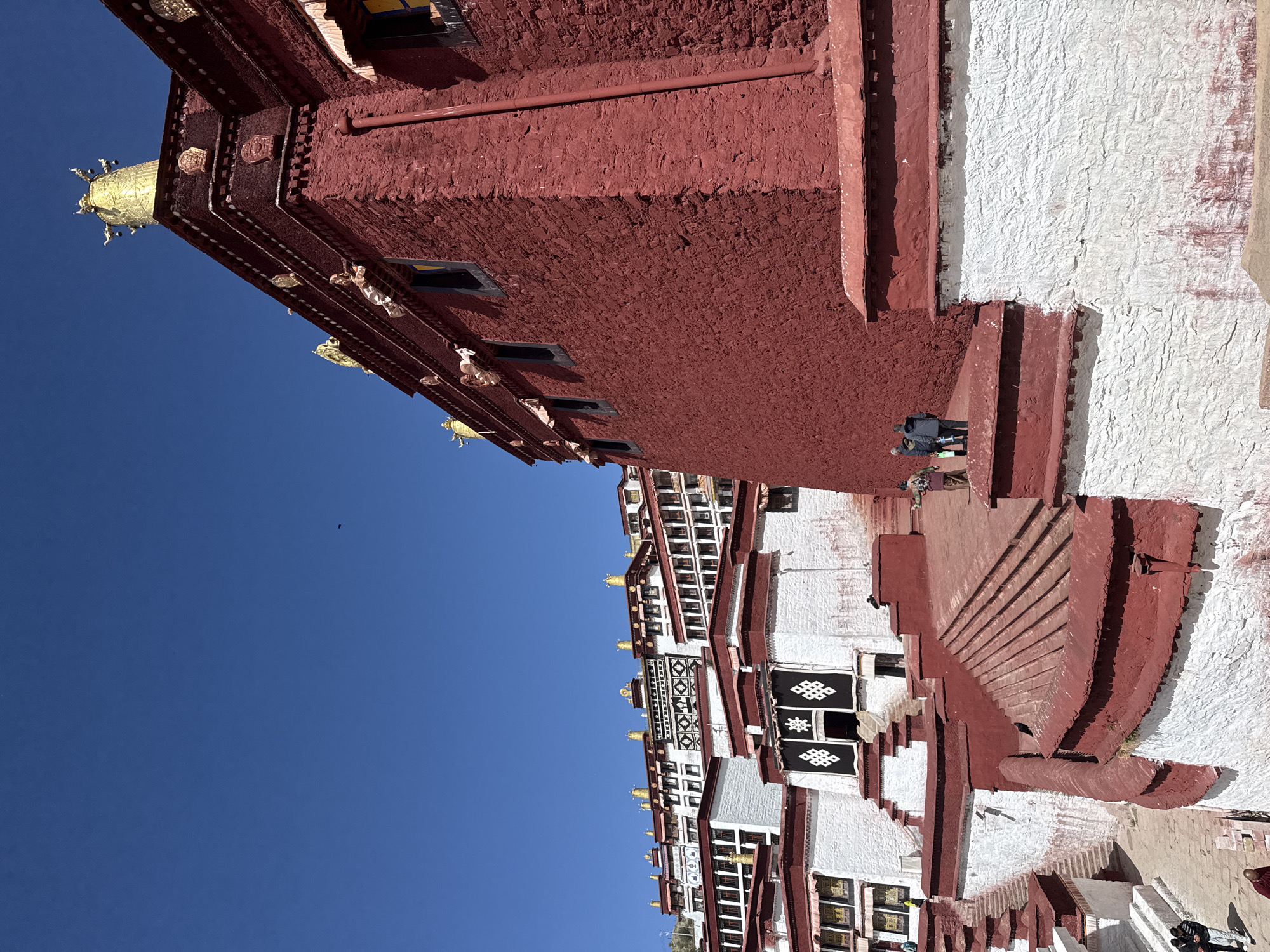 The image shows a building in Lhasa, China, specifically the Potala Palace. It is a large structure with red and white walls, and gold ornamentation. The lower section is predominantly white with red trim and details, including decorative windows and doorways. A set of stairs leads up the side of the building. Some people can be seen on the stairs. The sky is a clear, bright blue.