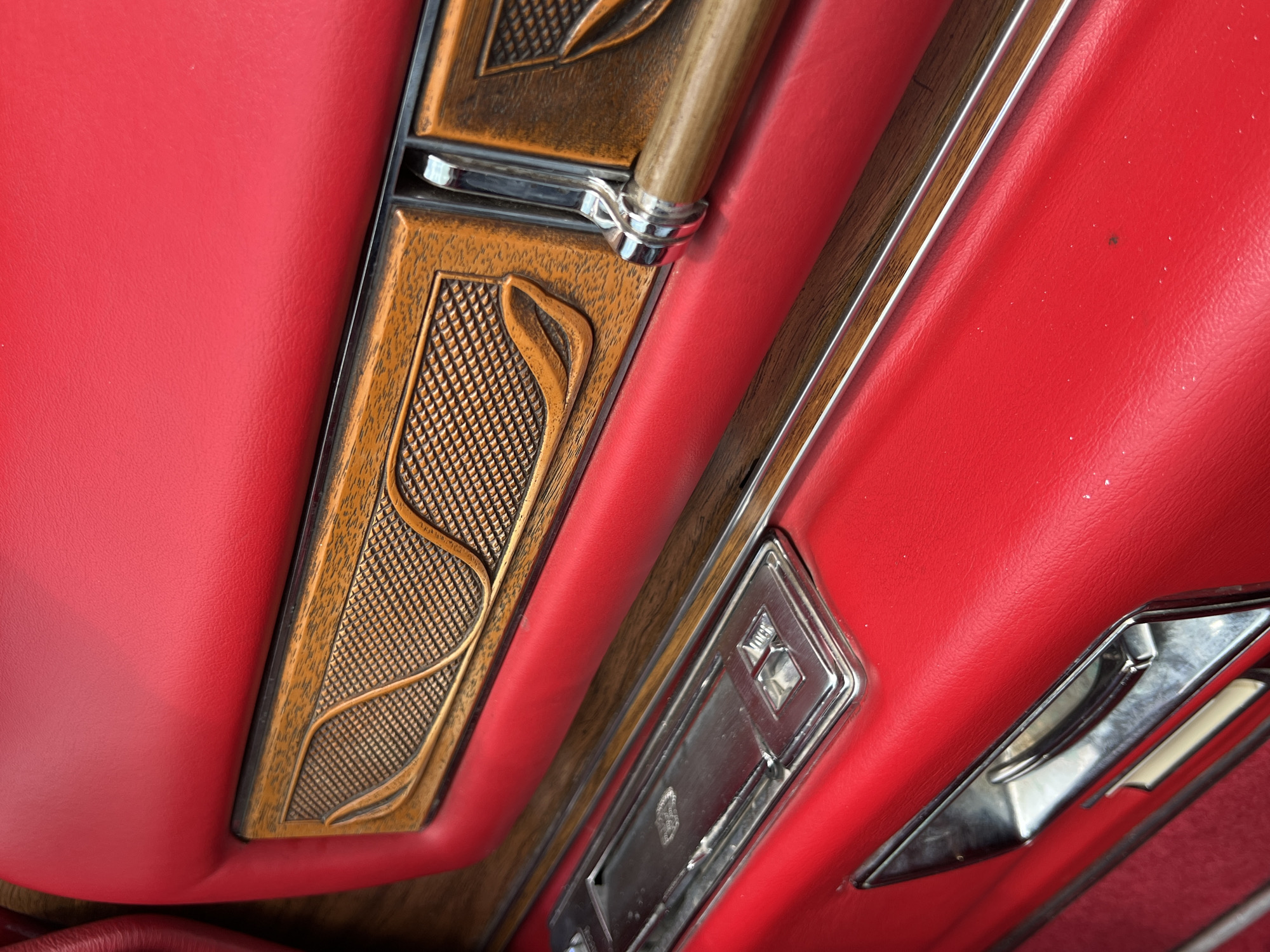 The image shows the interior of a classic car. The upholstery is red and appears to be leather or vinyl. There are woodgrain accents with silver trim. There is a handle and an ashtray, both with chrome detailing. The location is not discernible from the image.
