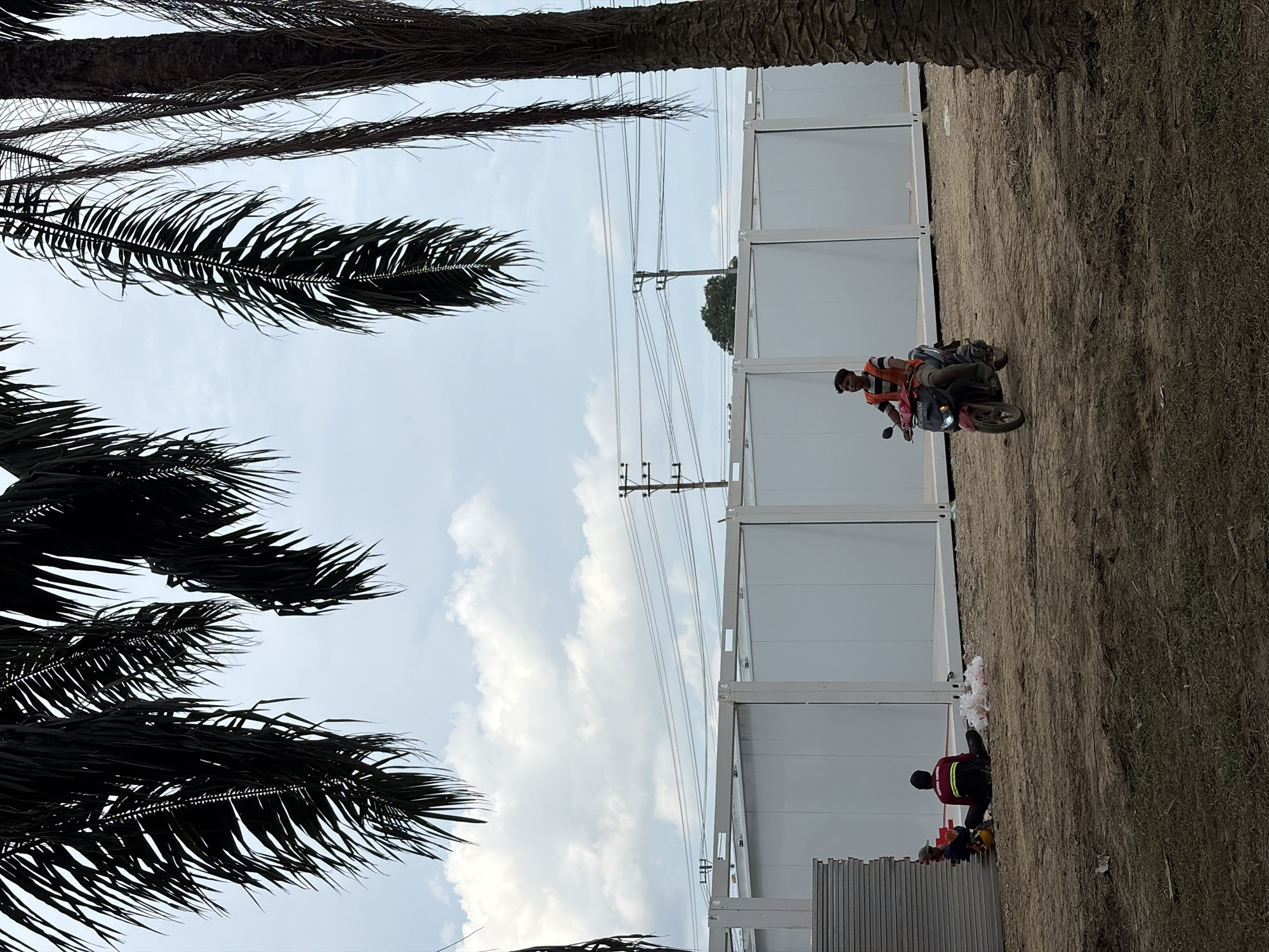 The image shows a construction site in Simpangempat, Indonesia, featuring white modular buildings, palm trees, and overhead power lines against a partly cloudy sky. A man wearing a striped shirt is seated on a motorcycle next to the buildings. Another man, clad in a red jacket with yellow stripes, appears to be working on the ground, near a stack of grey panels. The ground surface consists of dry dirt.