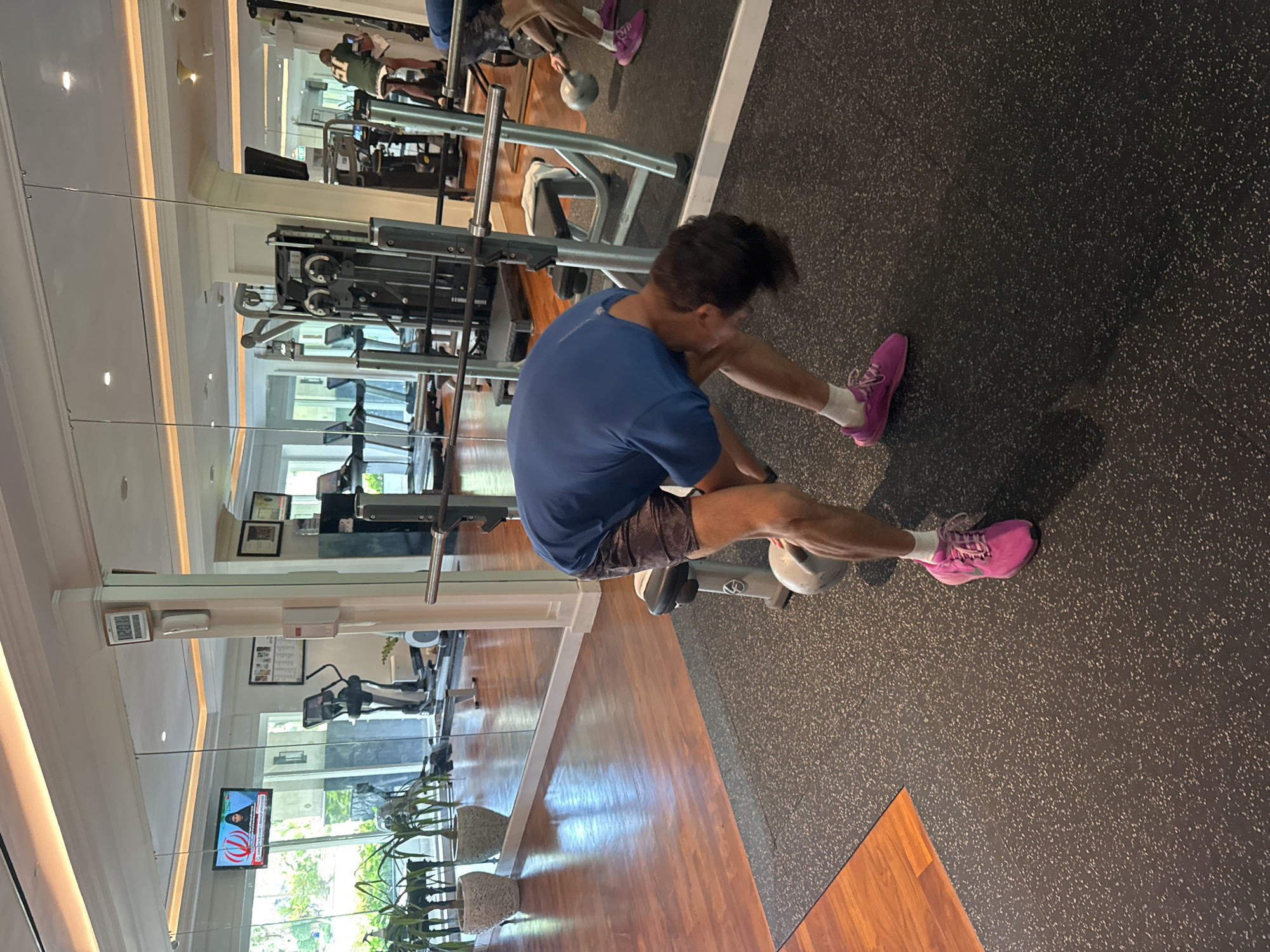 The image depicts a man sitting on a weight machine in a gym, likely in Kuta Selatan, Indonesia, although there is no definitive proof. The man is wearing a blue t-shirt, patterned shorts, white socks, and bright pink running shoes. He appears to be adjusting something on the machine. The gym features weight machines, treadmills, and other exercise equipment. The flooring is a combination of dark rubber and light-colored wood. Mirrors line the wall, reflecting the gym equipment and a television showing news. The gym appears well-lit and maintained.