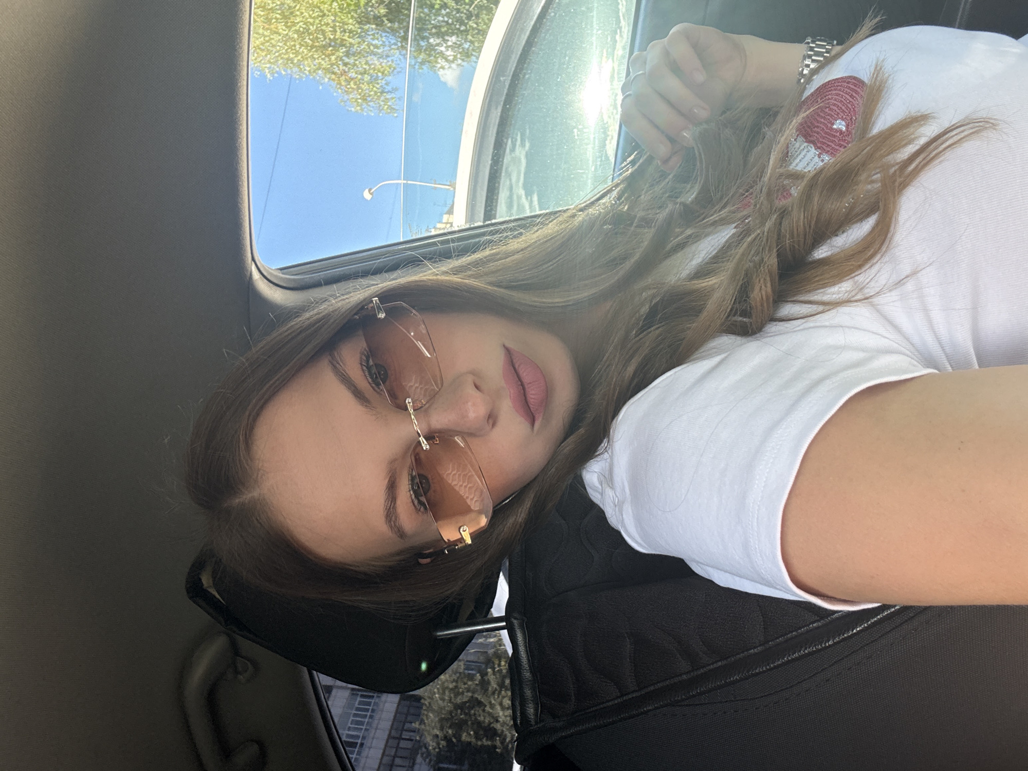 In a selfie taken from inside a vehicle, a young woman with long, light brown hair is seated in the driver's seat. She is wearing a white t-shirt and stylish rimless sunglasses with a subtle rose tint. Her left hand rests on her hair, and she is looking directly at the camera with a slight pout. Sunlight streams in from the right, casting a soft glow on her face and hair. The interior of the car is visible, with the black leather headrest of the passenger seat to her left. Outside the car window, a bright blue sky is visible with some trees and buildings in the background. The scene suggests a sunny day in an urban environment. The presence of a lamppost and the architectural style of the building in the background are consistent with a location in the city of Perm, Russia.