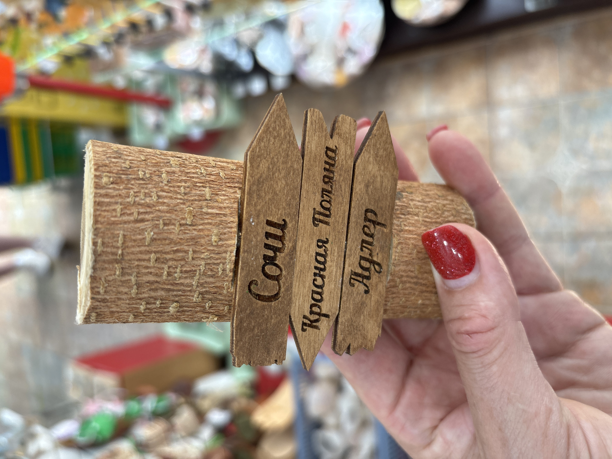 A close-up shot shows a wooden souvenir shaped like a log with three small, wooden signposts attached. The signs are carved with place names in Russian. The top sign reads "Сочи" (Sochi). The middle sign, pointing to the left, reads "Красная Поляна" (Krasnaya Polyana). The bottom sign, also pointing to the left, reads "Адлер" (Adler). A hand with red nail polish is holding the souvenir. The background is blurred, showing what appears to be a market or shop with shelves of goods and a person walking in the distance.