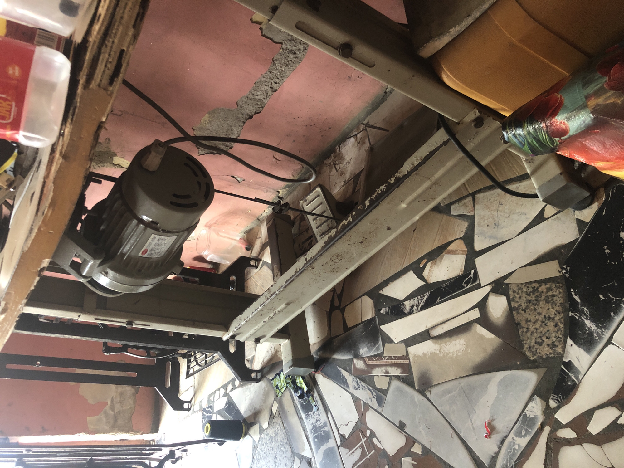 This image provides an under-table view of a sewing machine setup, likely within a workshop or home-based business in Ado, Nigeria. The primary subject is a large, grey industrial sewing machine motor, situated on the left, with a black power cable extending from it. A white label with unreadable technical text and numbers is affixed to the motor.
The machine rests on a sturdy metal frame, which supports a wooden table top, visible along the top edge of the image. The table and frame show signs of extensive use and wear. In the upper left, a small, clear plastic bottle with a red label displaying the partial word "JIK" is visible on the table.
The background features a worn-out, pink-painted ceiling or upper wall, with patches of plaster missing, revealing rough concrete beneath. To the right, parts of a yellow or orange plastic chair seat and a colorful, patterned bag or fabric are visible. The floor beneath the machine is distinct, created from a mosaic of broken tile pieces in various shades of white, black, grey, and brown. A small red piece, possibly a thread, lies on this mosaic floor. A black spool of thread with a yellow core is also visible on the lower left side, suggesting materials used in sewing.
No direct activity is taking place, but the arrangement indicates an active workspace. The general condition of the materials and surroundings suggests a practical, utilitarian environment.