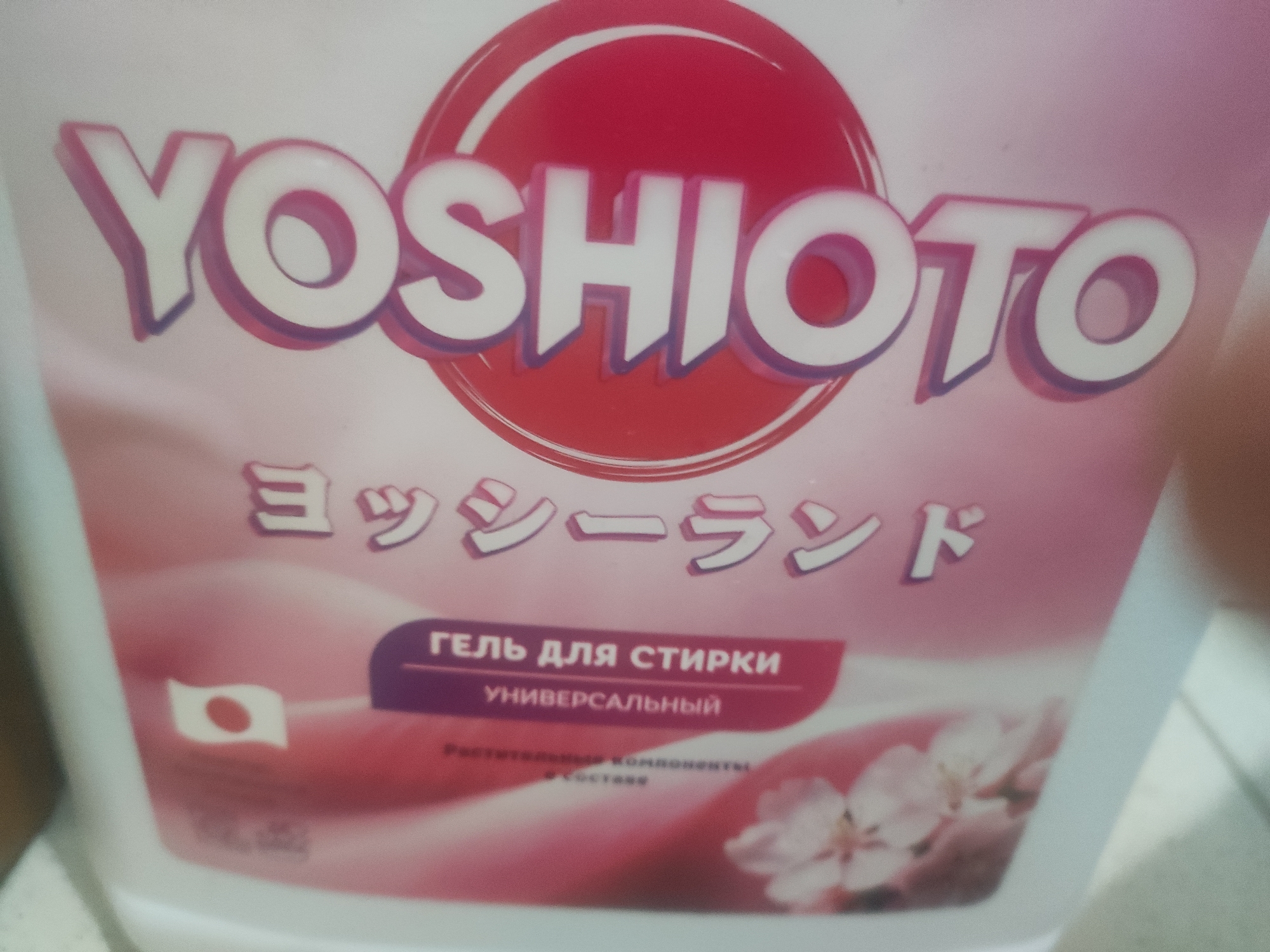 This is a close-up, eye-level, indoor shot of a white bottle with pink and red branding, likely a laundry detergent. The prominent text on the bottle reads "YOSHIOTO" in large white, stylized lettering with a purple outline. Below this, in smaller Japanese characters, is text that appears to say "ヨッシイランド" (Yoshii Land). A purple banner across the center displays Russian text that reads "ГЕЛЬ ДЛЯ СТИРКИ" (Gel for Washing) above "УНИВЕРСАЛЬНЫЙ" (Universal). To the left of the Russian text is a small image of the Japanese flag. The background is a soft pink gradient with an illustration of white and pink cherry blossoms at the bottom right. The overall presentation is clean and suggests a product marketed for its scent or origin. There are no people or discernible urban elements visible.