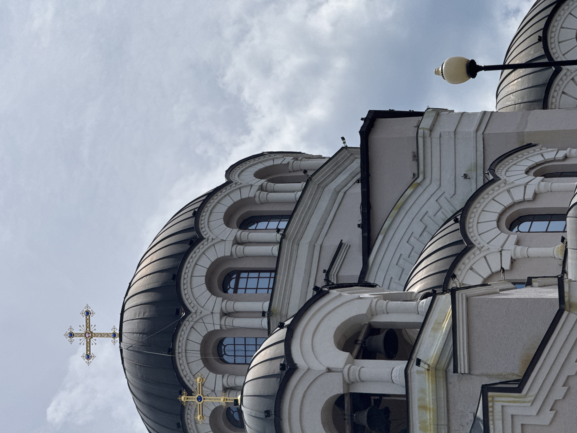 An upward-angled architectural shot shows a portion of a white building with dark domes, likely a church or cathedral. The building features rounded arches with columns and windows. The domes are striped in black and white, topped with gold crosses adorned with blue jewels. A black street lamp is visible on the right side of the frame. The sky is cloudy. The image is in Sochi, Russia.