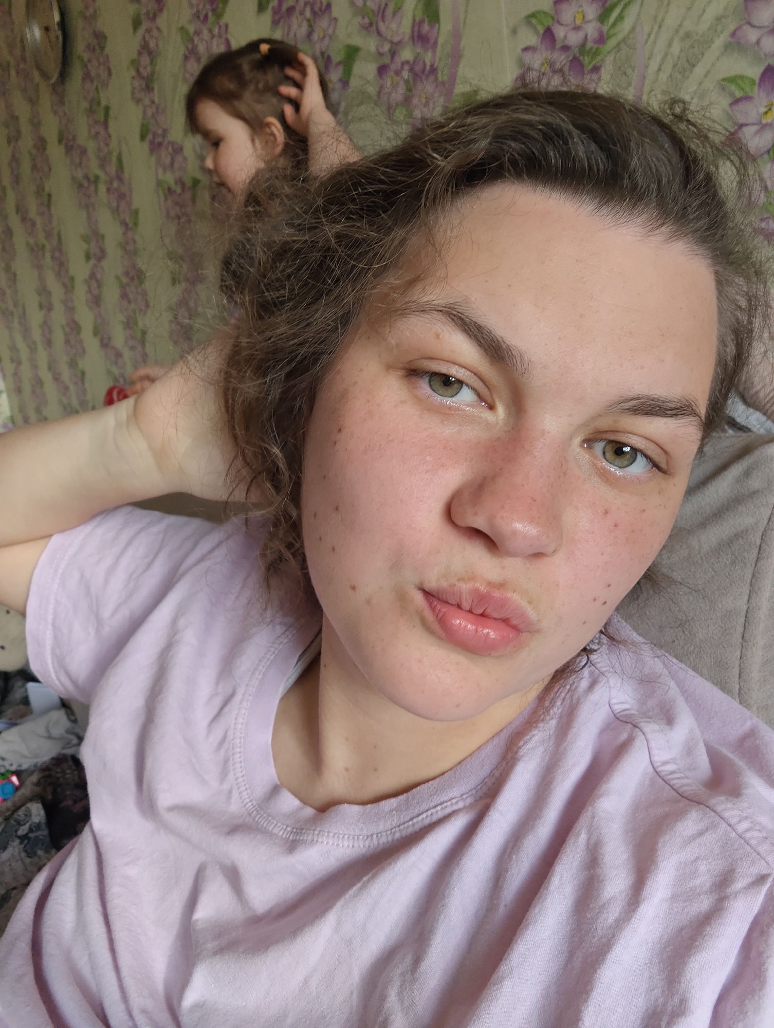 A close-up selfie shows a woman with light brown hair and green eyes, wearing a light purple T-shirt. Her right hand is raised near her head. She is pursing her lips. The background shows a young child and a floral patterned wall. The picture was likely taken indoors. The location is Уральское, Russia.