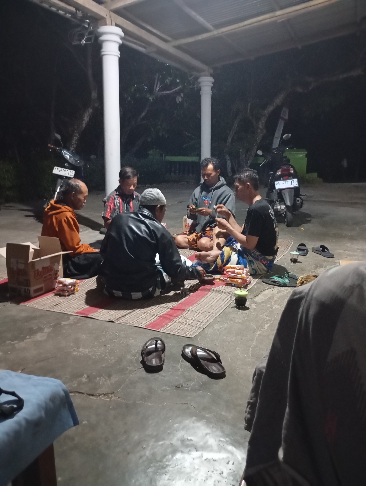 The image depicts a group of five men gathered on mats outdoors at night, likely in Wonogondo, Indonesia. The men are seated in a circle, engaged in a social gathering. One man in the foreground is wearing a black leather jacket and a white skullcap, and is seated with his legs crossed. To his left, another man in an orange jacket is also seated with his legs crossed. Two other men are seated in the background, one wearing a grey hoodie and the other in a checkered shirt. A fifth man is partially visible on the right side of the image, wearing a black t-shirt.
Various objects are scattered around the gathering. A cardboard box, a few packs of snacks, and some cups are placed on the mats. Two motorcycles are visible in the background, one parked to the left and another to the right. The scene is illuminated by artificial lighting, casting shadows on the concrete ground and the surrounding vegetation. The environment appears to be an open space with a covered structure supported by pillars, suggesting a patio or an outdoor seating area.