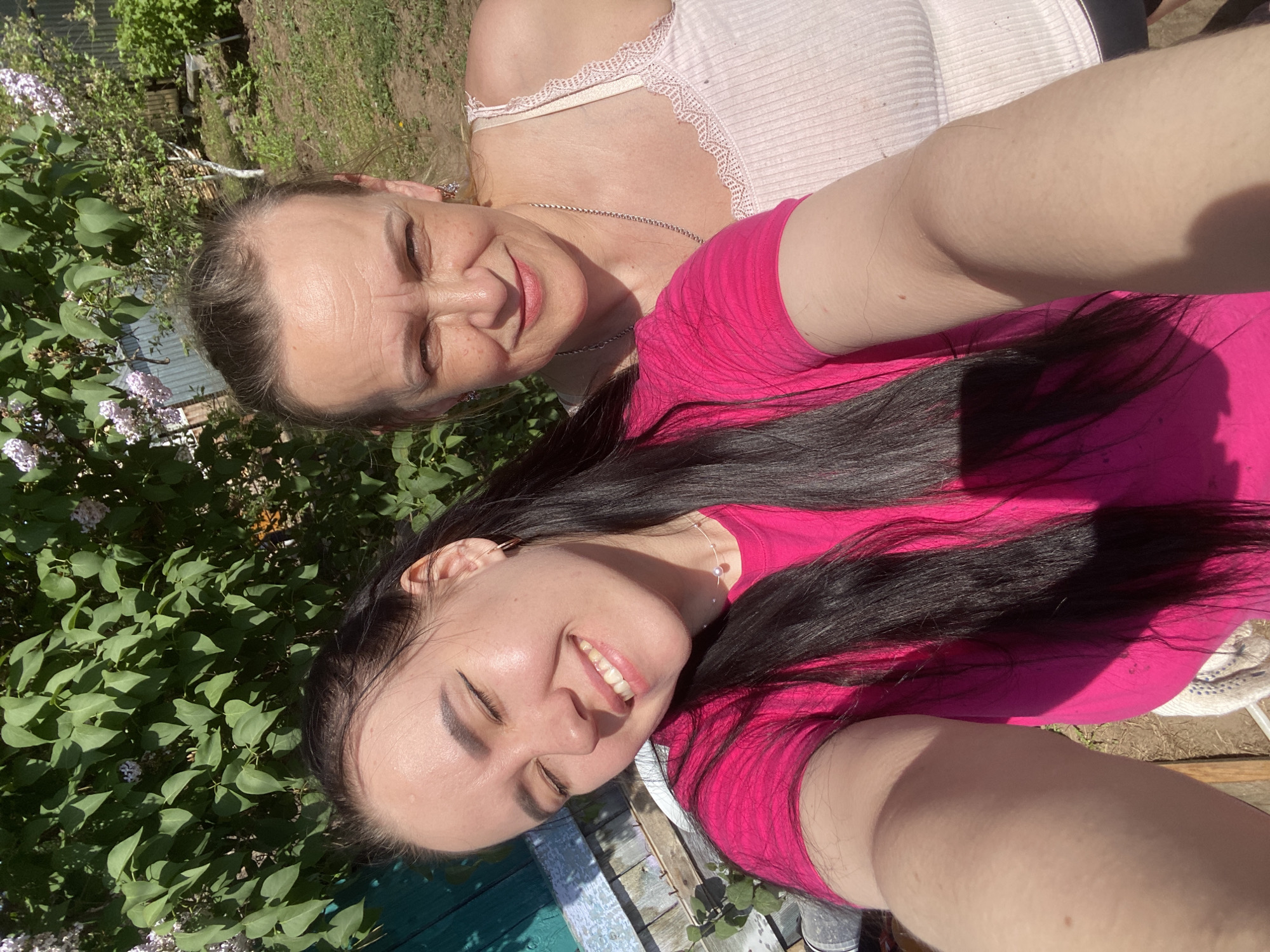 The image depicts two women in an outdoor setting. One woman, wearing a pink t-shirt and dark hair, smiles towards the camera. The other woman, slightly behind and above, has her eyes partially closed and also appears to smile. She is wearing a light-colored top with lace detailing. Lush green foliage is visible in the background, along with a few lilac flowers, suggesting a garden setting. The photo appears to have been taken in bright daylight. There is a wooden structure or surface on which the women may be positioned. While not explicit, the image is likely taken in a garden or park in the city of Chaikovsky, Russia, due to its location.