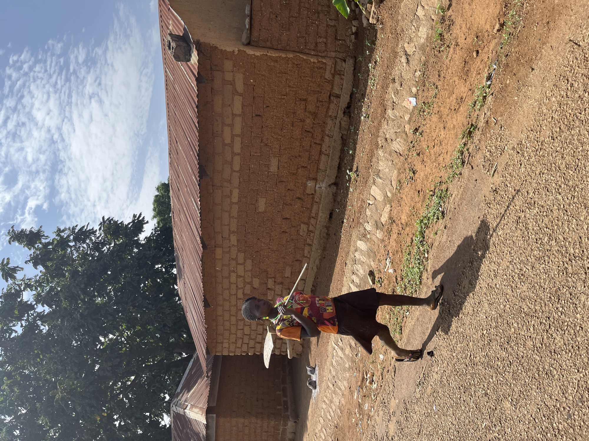 A young person stands on a dirt road in Barakin Momo Dogo, Nigeria, carrying a hoe and a stick. They wear a patterned shirt and a dark skirt, with sandals on their feet. Beside them is a building constructed of reddish-brown bricks, topped with a corrugated metal roof. A large tree is visible nearby. The sky is light blue with scattered white clouds. The road surface is uneven and appears to be a mix of dirt and small stones.