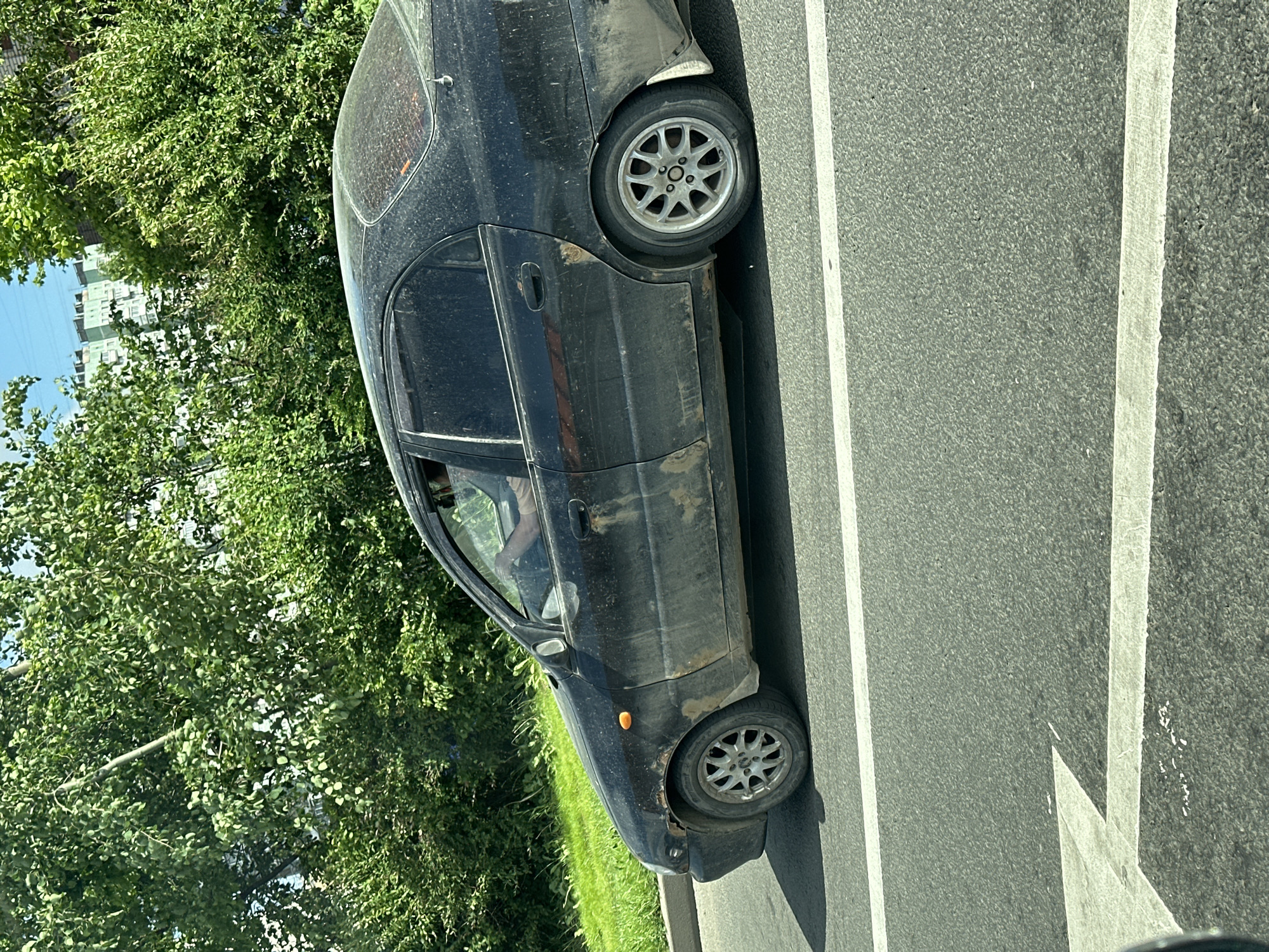 A black, heavily soiled sedan is driving down a paved road. The car has several areas of rust, particularly on the front fender and rear door panel. Tinted windows obscure the interior, but a person's arm is visible resting on the steering wheel. The car is moving from left to right in the frame, passing a grassy area and a line of trees. In the background, apartment buildings can be seen peeking through the foliage. The road has white lane markings, including a directional arrow painted on the asphalt. The sky is clear and blue. The overall impression is that of a vehicle driving through a suburban or semi-urban environment on a sunny day.