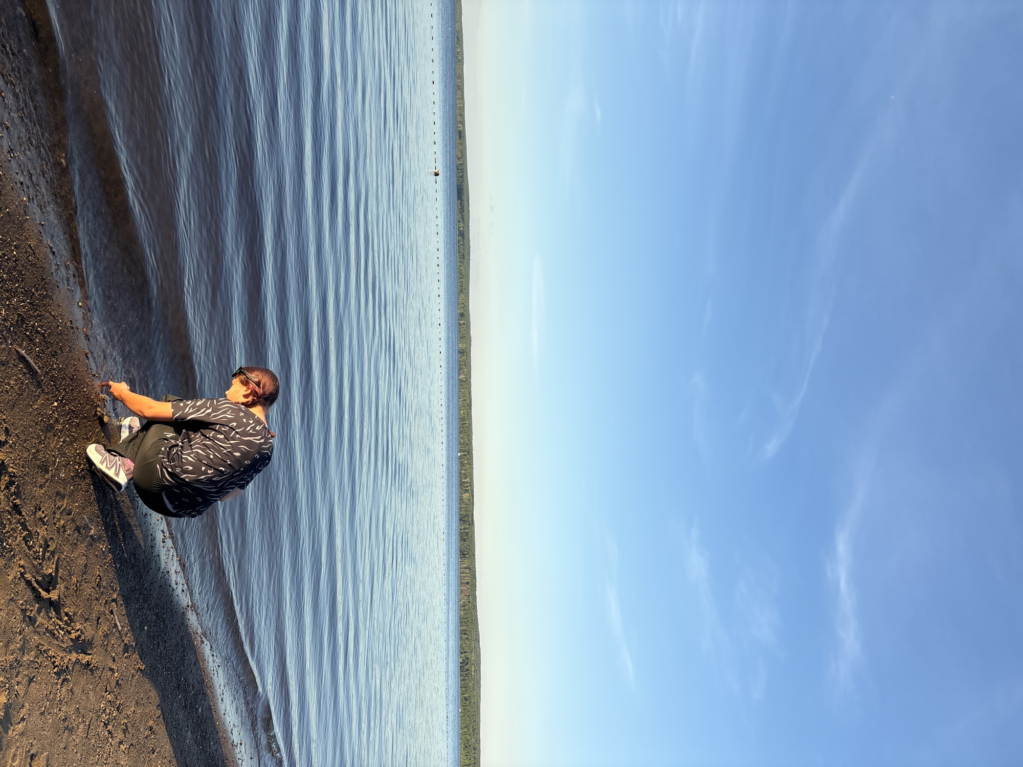The image depicts a person squatting by the shore of a lake, with a wooded shoreline in the distance. The person is wearing a dark patterned shirt, dark pants, and athletic shoes. They are leaning forward, appearing to interact with the water or the ground at the water's edge. The lake is calm with gentle ripples, reflecting the clear blue sky with wispy clouds. A line of buoys is visible across the water. The foreground consists of a sandy and pebbly shore. The overall scene suggests a serene outdoor setting, possibly a beach or lakeside area. The location is Chaikovsky, Russia.
