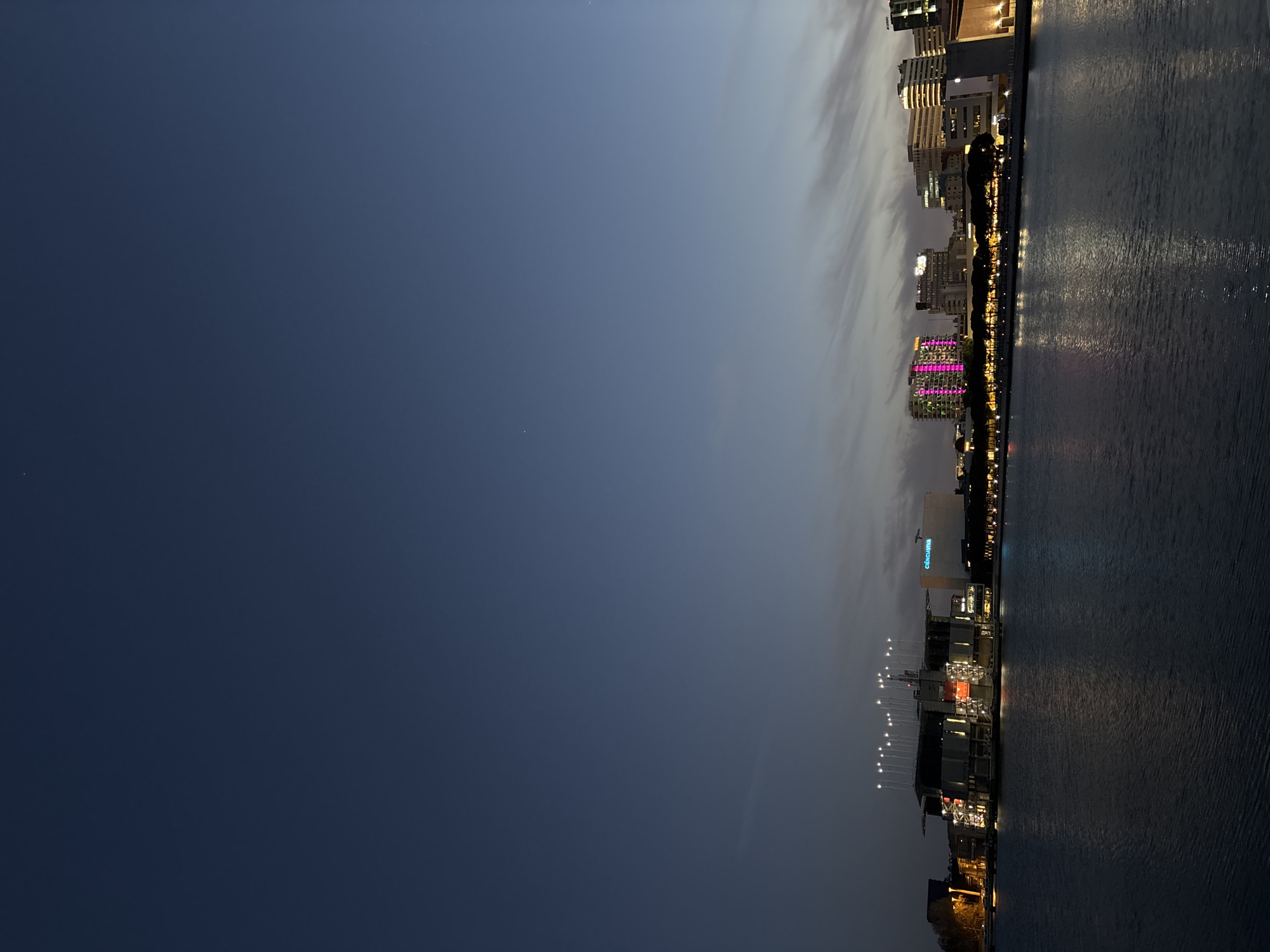 The image depicts the cityscape of Lisbon, Portugal, at dusk, situated along the waterfront. The view is likely taken from across the Tagus River, capturing a segment of the city's skyline. Buildings of varying heights, illuminated with artificial lights, punctuate the scene, with a notable structure displaying a pink facade. The water reflects the lights from the buildings, creating a shimmering effect. The sky transitions from a deep blue to a lighter shade toward the horizon.