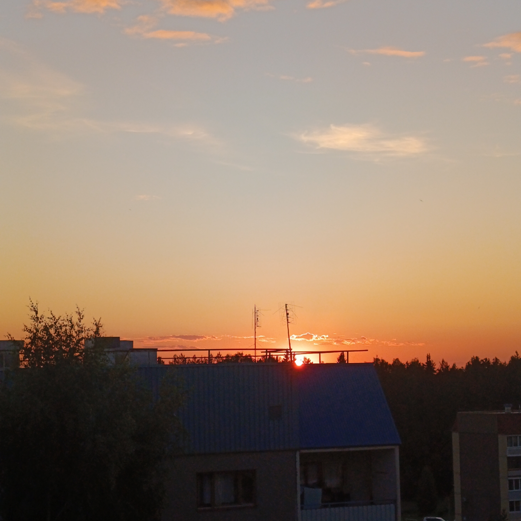 The image depicts a sunset over the city of Марковский, Russia, with an urban landscape in the foreground transitioning to a dense forest at the horizon. The sky exhibits a gradient, fading from light blue and pale orange at the top to intense orange and red hues near the horizon. Scattered, elongated clouds are present, some illuminated from below by the setting sun, particularly in the lower sky.
The sun, a bright red-orange disc, is partially obscured by thin clouds and the distant treeline, positioned slightly right of the vertical center axis. Its intense light creates a halo effect around itself and highlights the edges of nearby clouds.
In the midground, a two-story building with a dark blue corrugated metal roof and grey-painted lower walls is prominent. The top edge of this building features a dark railing, behind which two distinct television or radio antennas are silhouetted against the bright sky. One antenna, on the left, is vertical with multiple cross elements, while the one on the right is angled upwards and also has a complex, multi-element structure. The lower part of this building shows two dark rectangular windows on the left and a recessed balcony area on the right, also in shadow. To the left of this building, a leafy deciduous tree with dark foliage is silhouetted.
To the far right, a portion of another multi-story building is visible, also silhouetted against the orange sky. This building appears to have a lighter, possibly reddish, upper section and multiple visible windows.
The background is dominated by a dark, dense coniferous forest that forms a continuous silhouette along the horizon line. No people or moving vehicles are discernibly visible in the frame.