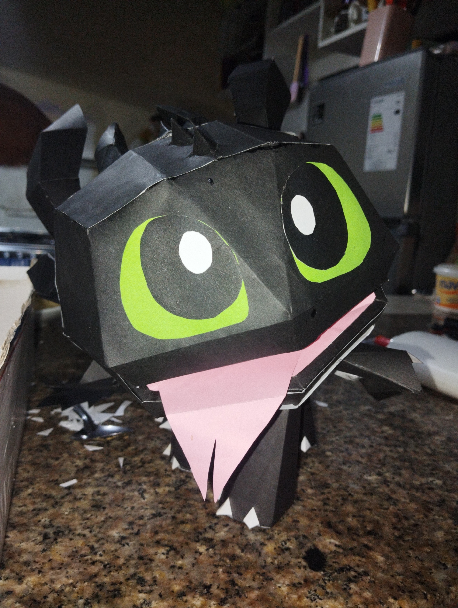 A papercraft model of Toothless, the dragon from the "How to Train Your Dragon" franchise, sits on a granite countertop. The model is primarily black with green and white eyes and a pink tongue. The background includes a refrigerator and scattered scraps of paper. The location is not discernible from the image.