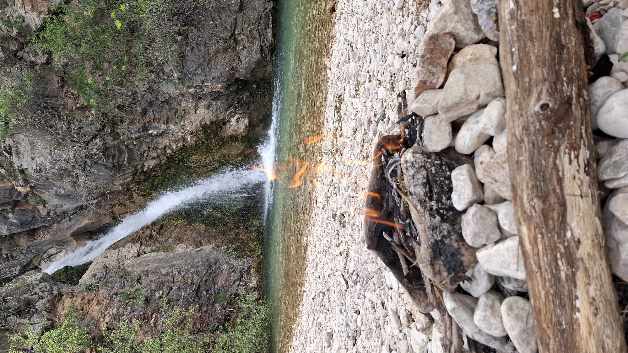 A long shot shows a waterfall flowing into a stream in what appears to be Boreti, Montenegro. To the right of the water, a small fire burns between rocks and a larger piece of wood. The camera angle is tilted, making the image appear as if it were turned on its side. The background is a rocky landscape with vegetation. The foreground is mostly covered with light-colored pebbles. The lighting suggests it's daytime.
