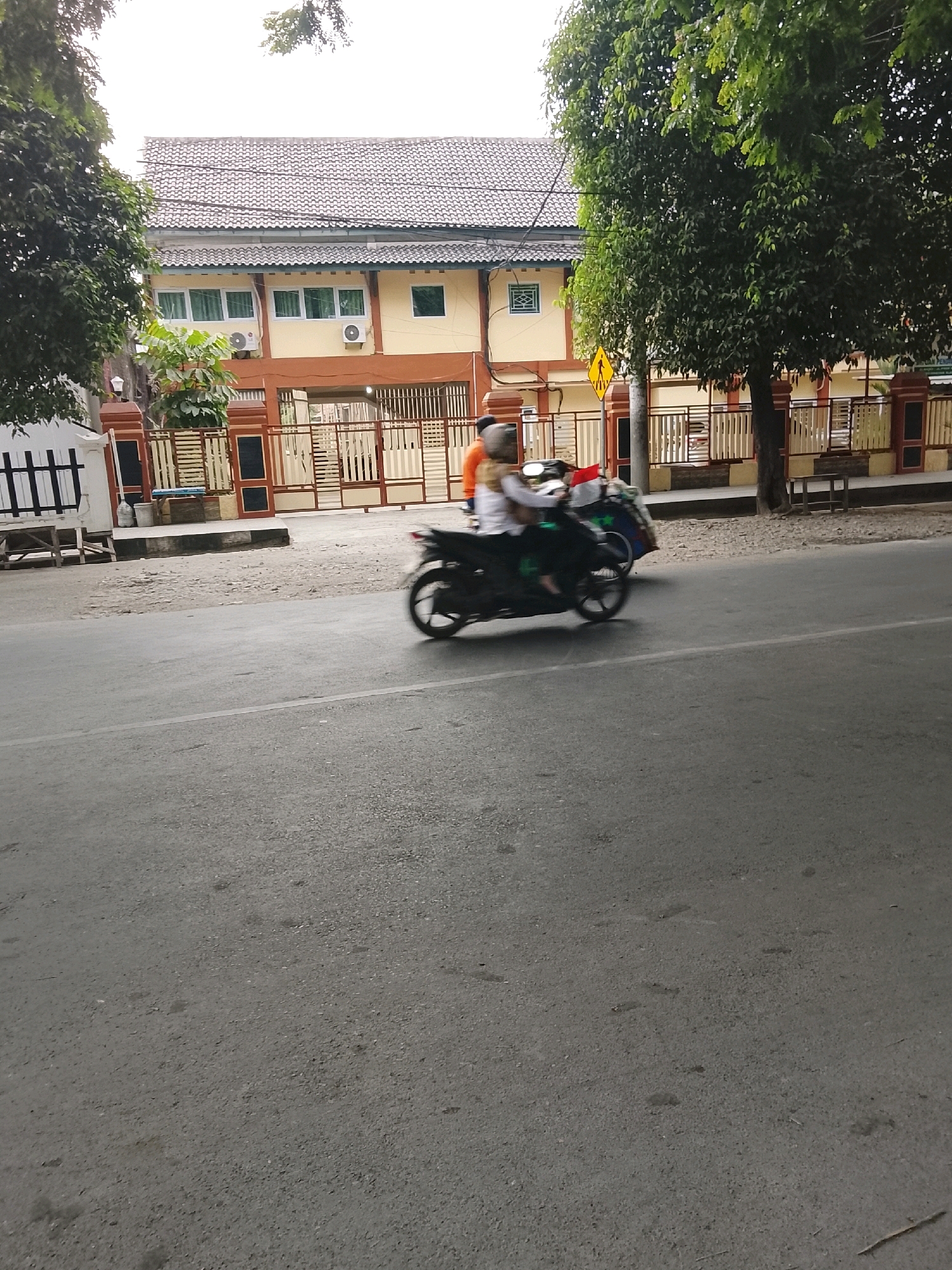 The image depicts a street scene in Slawi, Indonesia, with a building and trees in the background. A motorcycle carrying two individuals is moving on the road, creating motion blur. The building appears to be a two-story structure with a tiled roof and yellow walls. A fence with brown and white accents separates the building from the street. Green trees flank the building and are also visible on the left side of the image. The road is asphalt, with a white line marking the lane. The sky is overcast. A yellow pedestrian crossing sign is visible on the right side of the road.