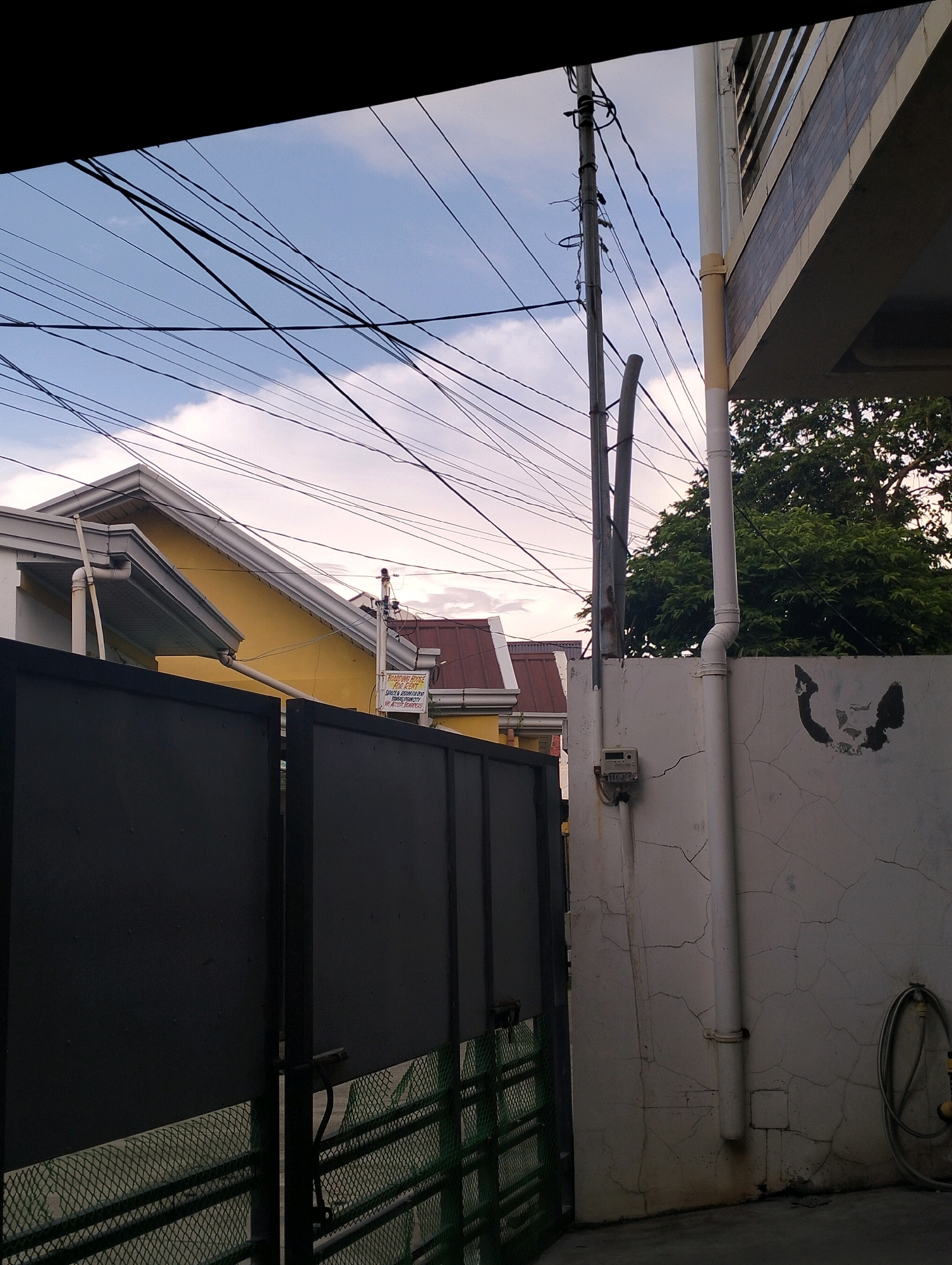 The image depicts an outdoor scene in Caoayan, Philippines, featuring a residential or commercial property. In the foreground, a dark, solid gate made of metal is visible, partially obscuring a green mesh fence underneath. To the right of the gate, a light-colored concrete wall is prominent, marked with cracks and featuring a white pipe running vertically down its surface. A hose coiled on the wall is also visible. Above the wall, a portion of a building with a white facade and a tiled roof can be seen. Overhead, a network of electrical wires crisscrosses a pale blue sky with scattered clouds. In the middle ground, a yellow building with a white-trimmed roof and a smaller building with a red tile roof are situated behind the gate. A utility pole stands to the right of the yellow building, supporting the electrical wires. A sign is affixed to the yellow building, though the text is not entirely legible. The overall impression is a somewhat cluttered urban environment, common in residential areas of the Philippines.
