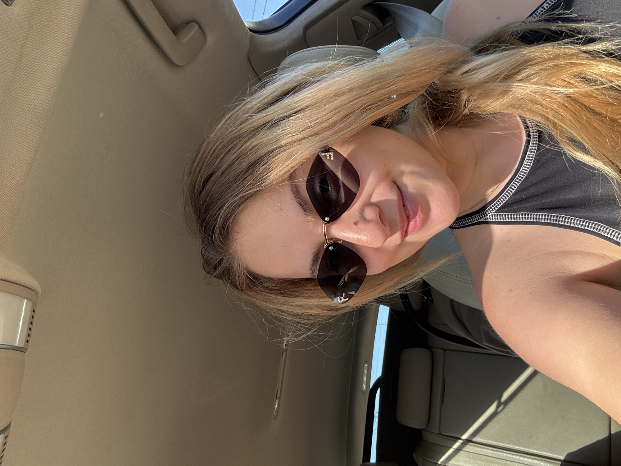 A young Caucasian woman with blonde hair and sunglasses is sitting in the passenger seat of a car. She is smiling and looking at the camera. The interior of the car is visible, including the seats and the ceiling. Sunlight is streaming in through the windows, casting shadows on the seats. The woman is wearing a black tank top. The scene appears to be during the daytime, possibly in a suburban or rural setting.