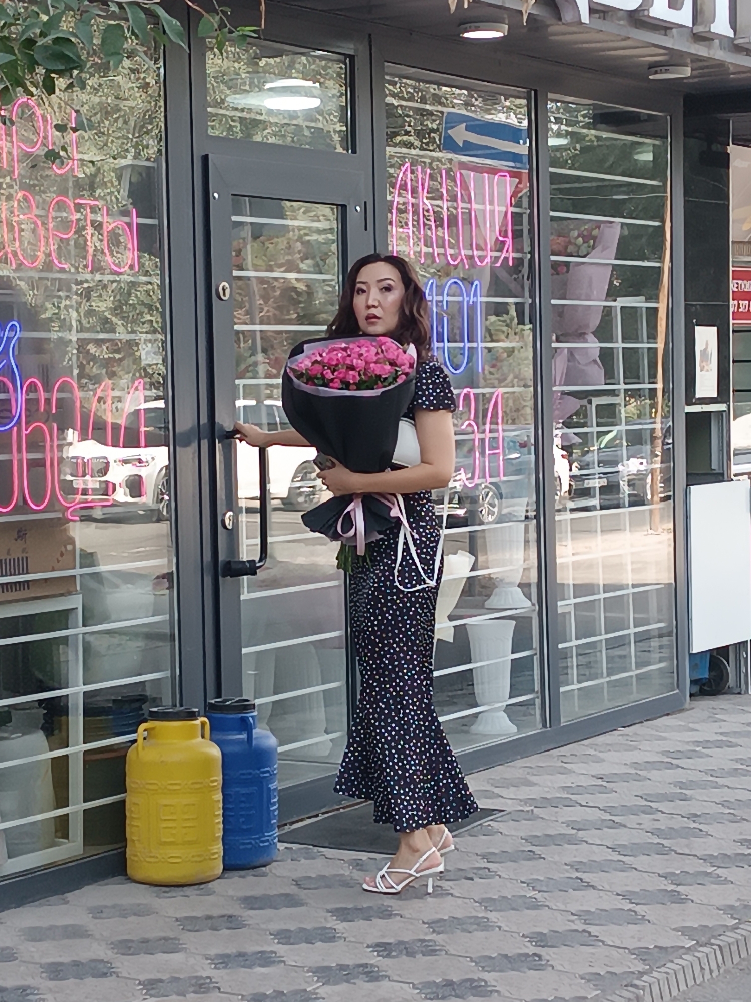 A woman in a black and white polka dot dress and white heeled sandals is standing outside a shop in Bishkek, Kyrgyzstan. She is holding a large bouquet of pink roses and a smartphone. The shop has large glass windows with neon signs in Cyrillic. In front of the shop are two large plastic containers, one yellow and one blue.