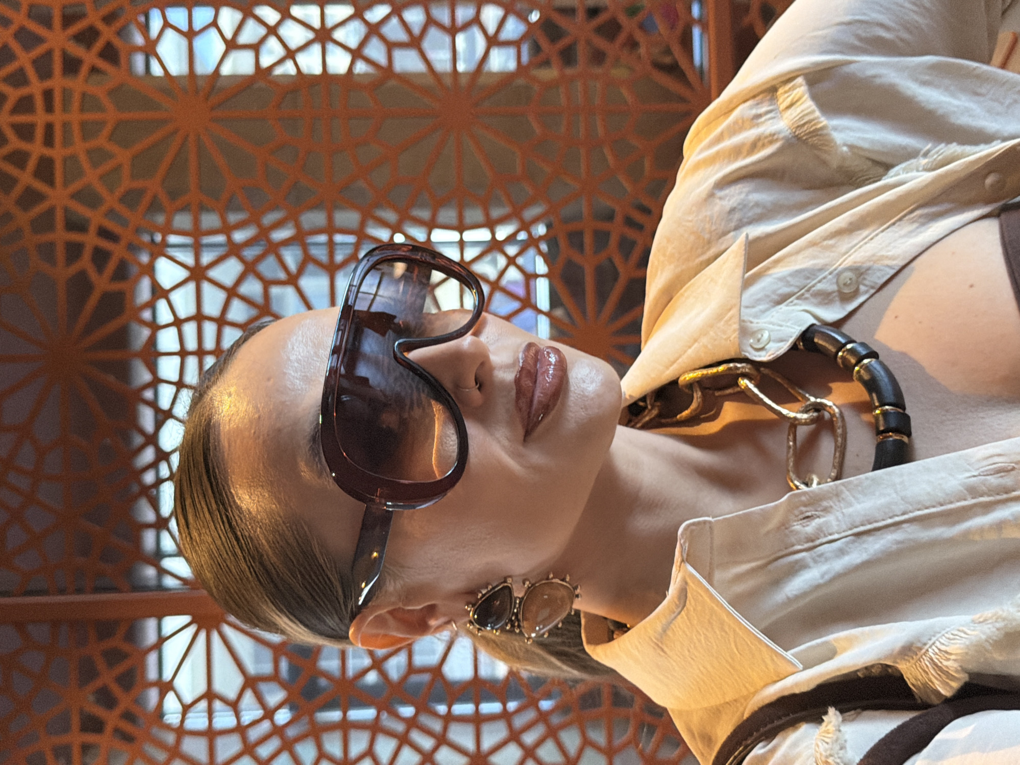 A woman with slicked-back hair is wearing oversized sunglasses and a light-colored shirt with an elaborate necklace and earrings. She is positioned in front of an orange, geometric screen that appears to be an architectural element, possibly a facade. Based on the metadata, the photograph was taken in Krasnoyarsk, Russia. The exact location within the city is not specified, but the presence of the patterned screen suggests a location with modern architectural design. The subject is looking slightly upwards, and the camera angle is slightly low, giving the impression that the photo was taken from a slightly lower position. The lighting seems bright, emphasizing the textures of the clothing and jewelry.
