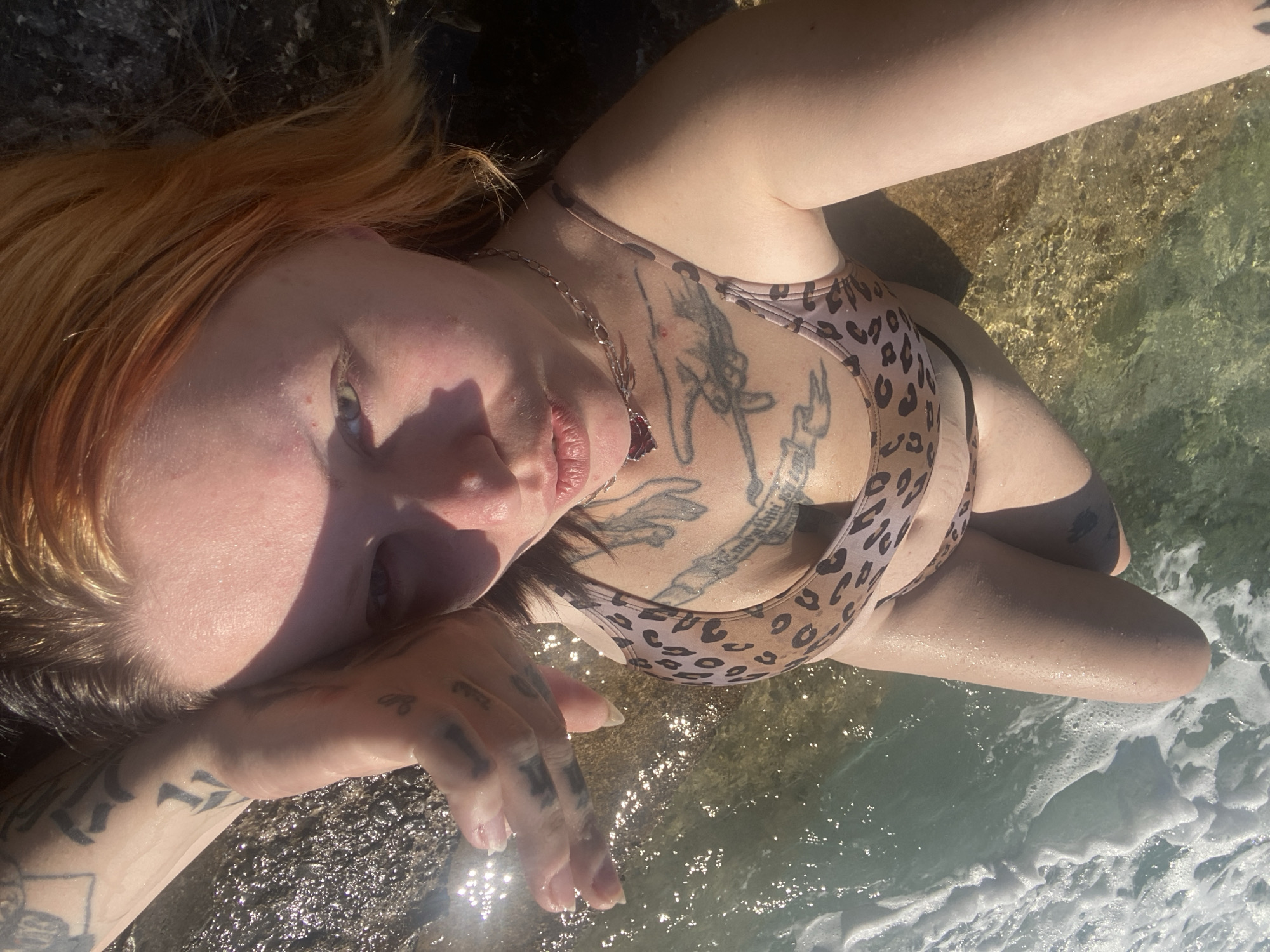 A woman with fair skin and orange-toned hair is lying on a rocky surface near water, presumably in Bar, Montenegro, though the specific location is not identifiable from the image alone. She is wearing a leopard-print bikini. Multiple tattoos are visible on her chest, arms, and hands. Her left hand is raised and touching the water. The water appears clear and shallow, with some white foamy waves visible. The overall lighting suggests a sunny day.