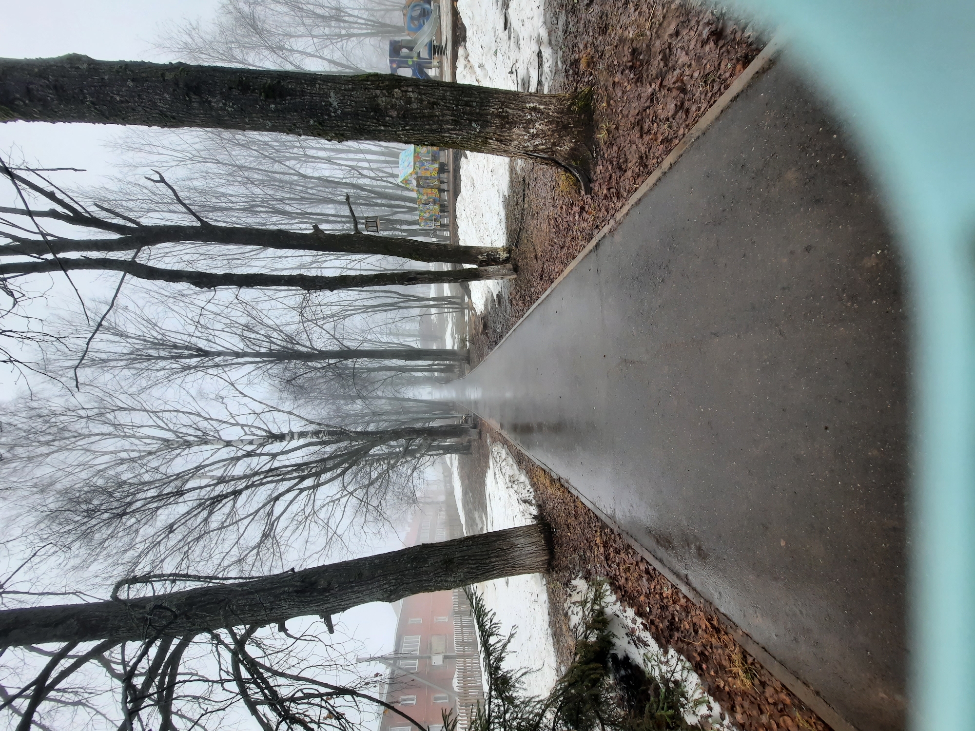 The image depicts a wet, paved pathway lined with bare trees, characteristic of late fall or early spring. The pathway stretches into a hazy distance, suggesting fog or mist. Patches of melting snow are visible alongside the path, mixed with fallen leaves. In the background, a playground and a red brick building can be discerned, indicating the setting is likely a park or residential area. The lower right corner of the image is partially obscured by a blue-green object, perhaps part of a structure or foliage. The scene is indicative of a cold, damp day in a public space in Krasnogorskoye, Russia.