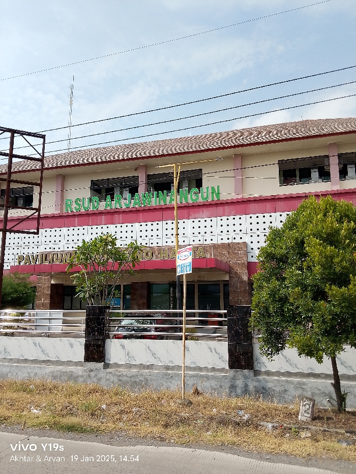 The image displays the exterior of a hospital building, identified as RSUD Arjawinangun, located in Arjawinangun, Indonesia. The scene is captured during daylight, likely in the afternoon, given the bright illumination and visible shadows. The sky is a clear blue with some scattered white clouds.
The hospital building is a multi-story structure with a distinctive brown tiled roof. Its facade features a light-colored upper section, a prominent red band, and a lower section with a white pattern of small black squares. Large green letters on the red band clearly spell "RSUD ARJAWINANGUN." Below this, above the main entrance, a darker band bears red text that reads "PAVILIUN SOEKARNO-HATTA." The entrance area itself has brown pillars and a glass facade.
In front of the hospital, there's a low fence or wall with a white, marble-patterned base and silver metal railings. Various trees and shrubs are planted both behind and in front of this fence, including a larger tree on the right side of the frame. A small blue and red sign, likely for an "Indomaret" convenience store, is attached to a pole near the entrance. The immediate foreground consists of dry grass and dirt, with a paved road visible at the very bottom. A small concrete marker with the number "2" is embedded in the ground on the right. Power lines stretch across the upper portion of the sky. A car can be faintly seen parked behind the fence.
The image carries a watermark indicating it was taken with a "vivo Y19s" by "Akhtar & Arvan" on "19 Jan 2025, 14.54."