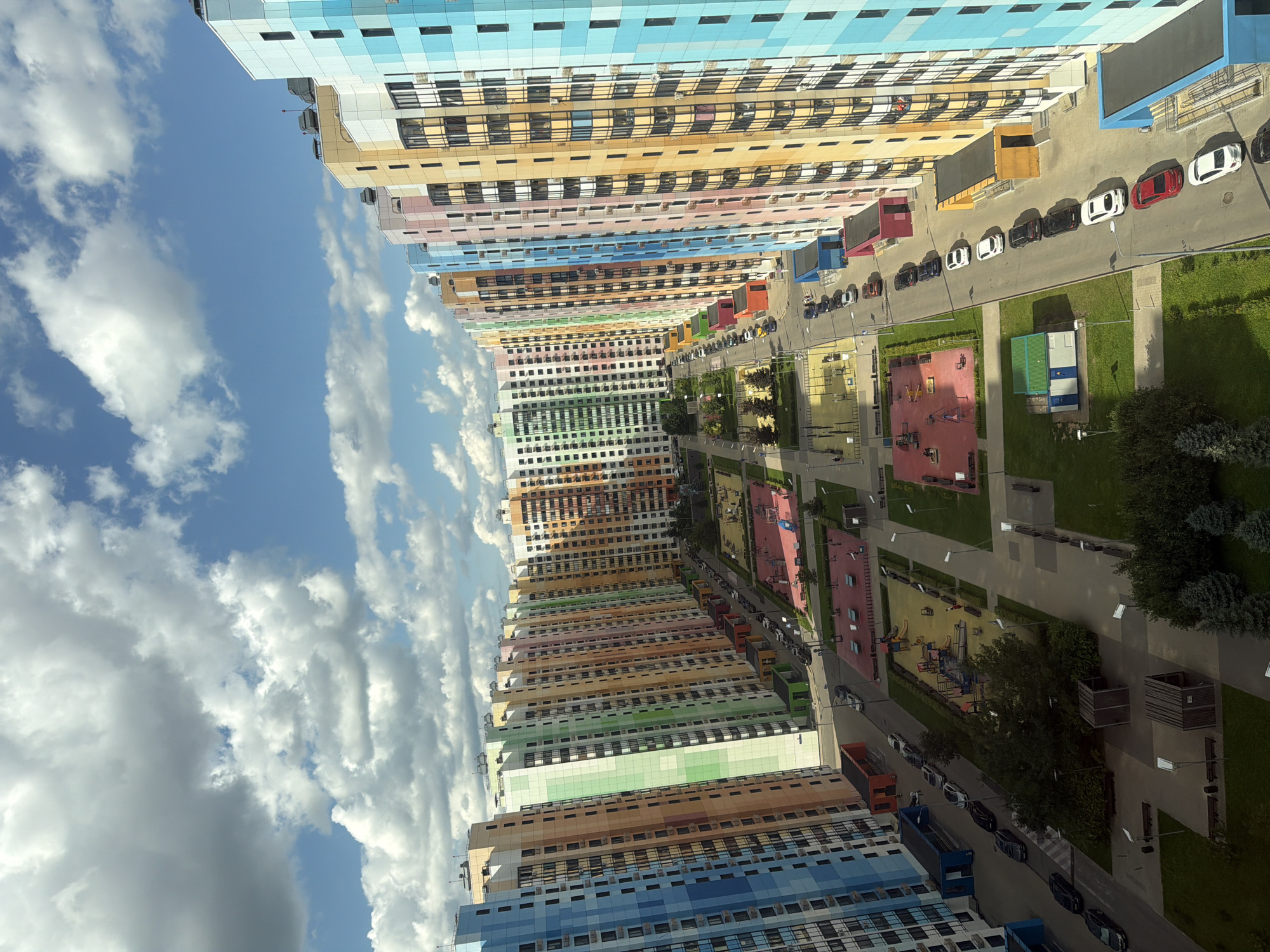 An aerial, high-angle shot captures a modern residential complex in the city of Unknown, Unknown. The scene is dominated by a row of tall, colorful apartment buildings, each featuring a distinctive facade with alternating vertical panels of blue, orange, yellow, pink, green, and white. These buildings are arranged in a curved formation, creating a courtyard-like space between them. Within this courtyard, well-maintained playgrounds with red and yellow equipment are visible, along with a paved walkway and green lawn areas. A street lined with parked cars runs parallel to the buildings. The sky above is a bright blue, dotted with fluffy white clouds, indicating a clear and sunny day.