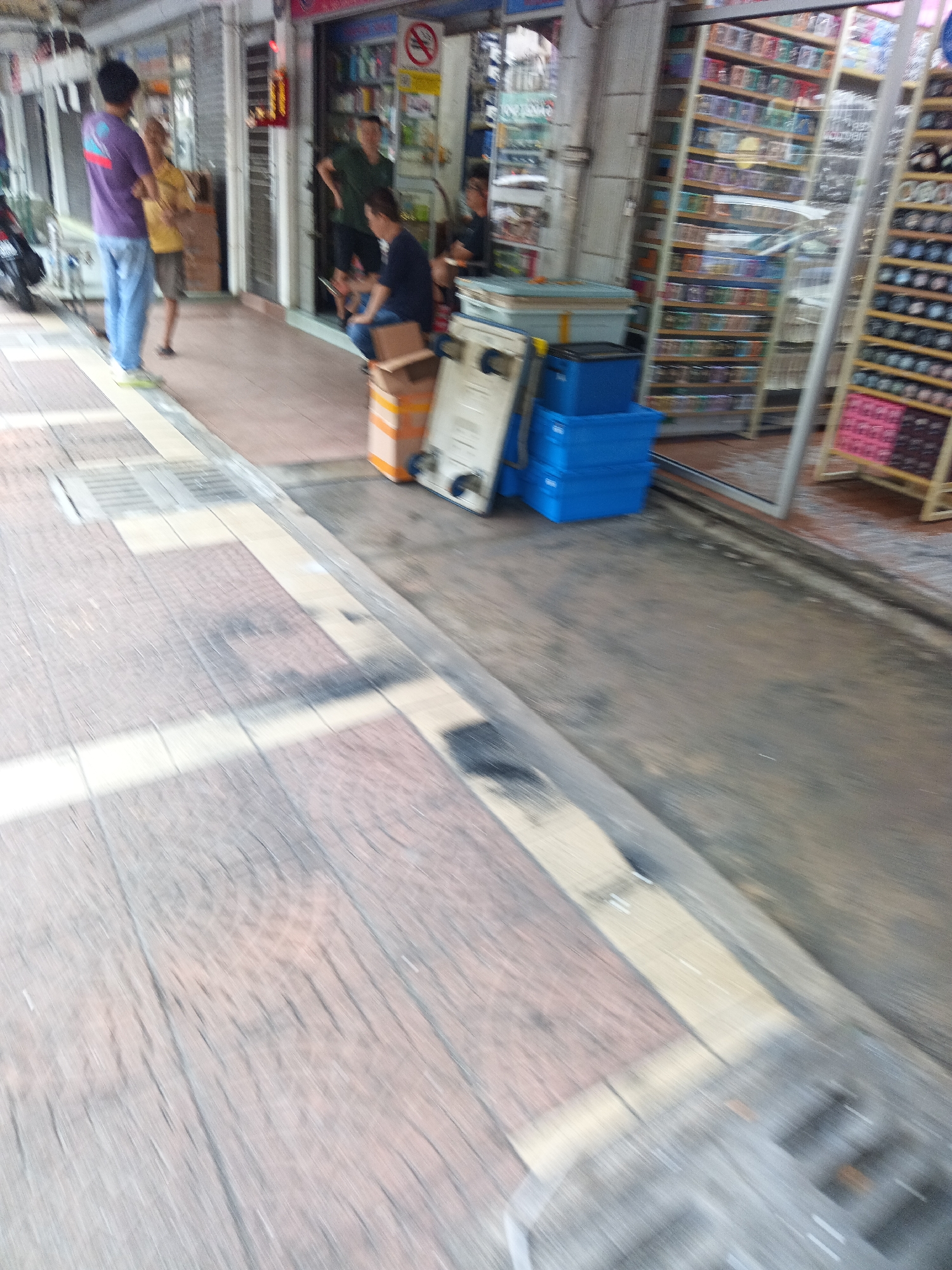 The image depicts a street scene in Kuala Lumpur, Malaysia, likely in a commercial area. The foreground shows a textured pavement, possibly a sidewalk, with white lines and grates. In the mid-ground, several people are visible. Two individuals stand near the entrance of a building, one wearing a purple t-shirt and blue pants, the other in a yellow shirt. Another person is seated outside a shop, wearing a dark blue shirt. In front of the shop, stacks of blue plastic containers and a cardboard box are placed on the ground. The background reveals the storefronts of shops, with shelves displaying merchandise and reflective surfaces suggesting glass doors or windows. The overall impression is that of a bustling urban environment.
