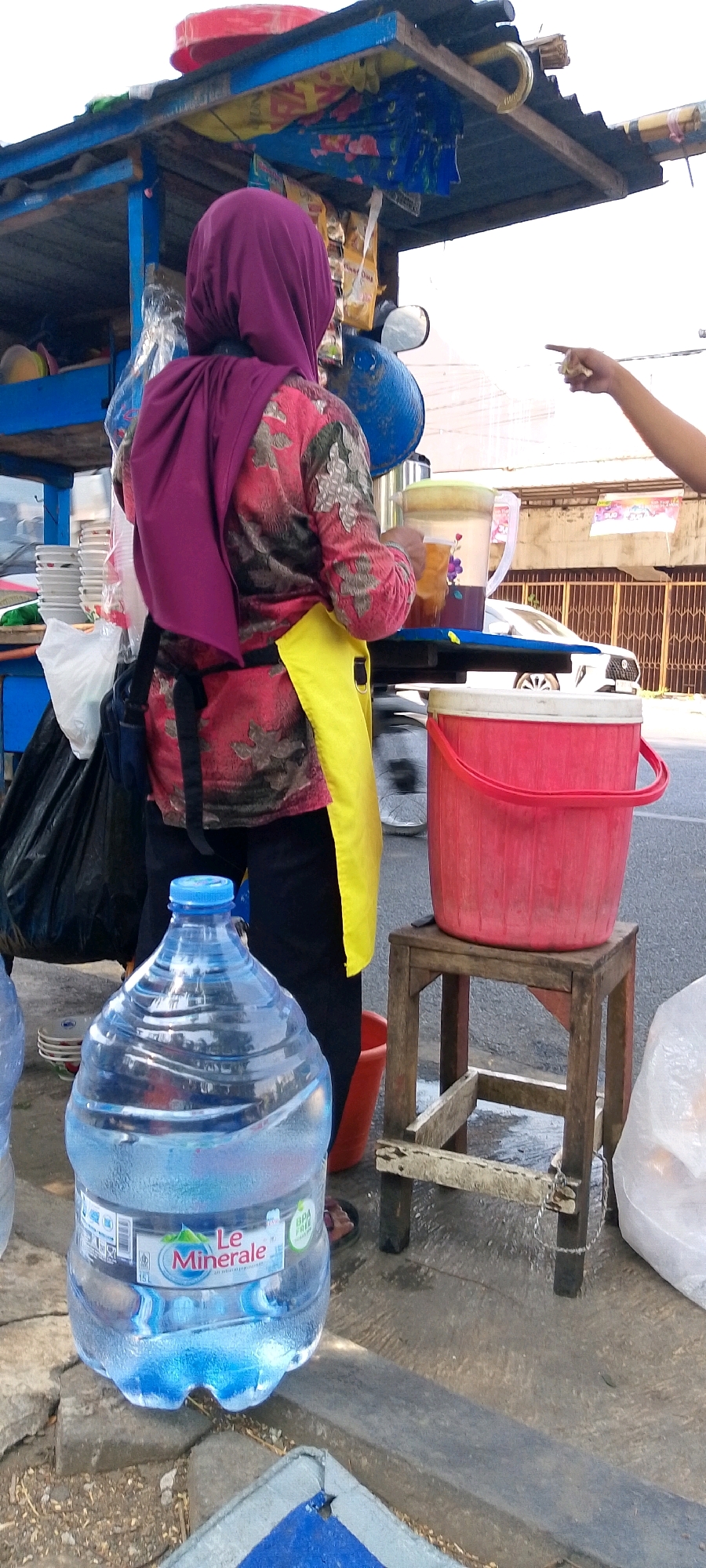 A person wearing a magenta hijab and a colorful patterned shirt with a yellow apron is standing at an outdoor food stall in Semarang, Indonesia. They are holding a clear plastic cup filled with a brownish liquid. In front of them, a large blue water cooler bottle is prominent. To their right, a red bucket sits on a small wooden stool. The stall itself is decorated with various items, including umbrellas. The ground appears to be paved, and a white car is visible in the background, suggesting a street setting. The overall scene depicts a typical day at a street-side vendor in Indonesia.