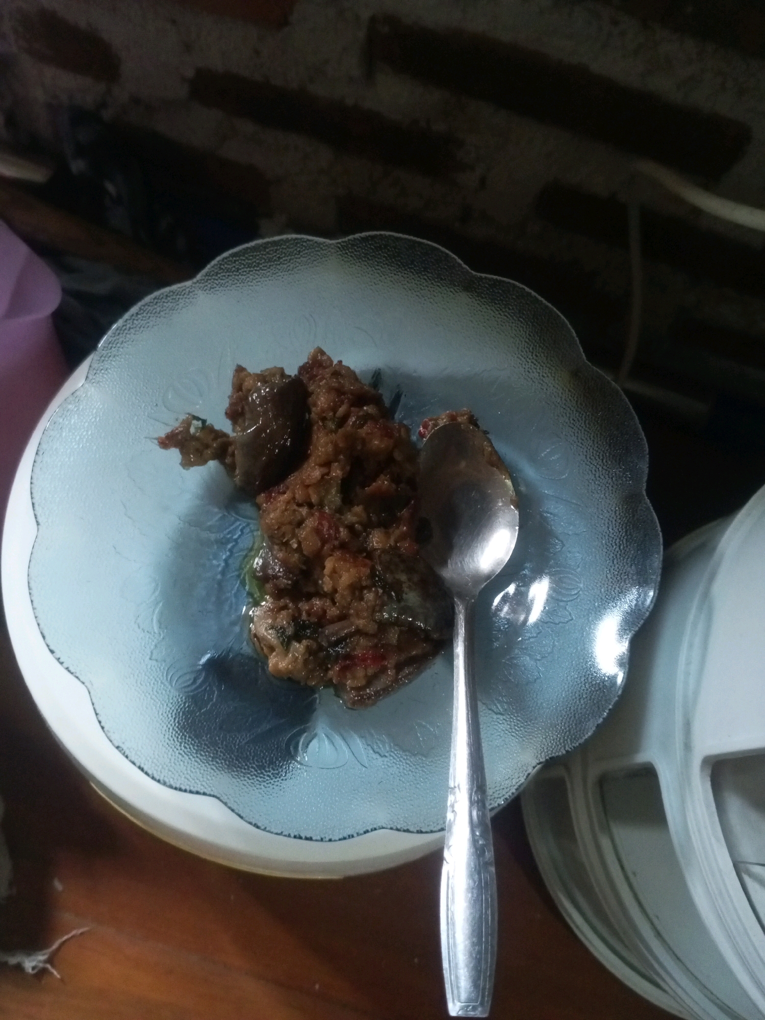 A close-up, overhead shot shows a serving of Indonesian cuisine on a patterned glass plate. The food appears to be a spicy, rich mixture, possibly a stew or stir-fry, with visible chunks of meat, vegetables, and chili peppers. A silver spoon rests on the right side of the plate, slightly embedded in the food. The plate is clear with a wavy, scalloped edge and a textured surface featuring embossed floral patterns. It sits atop a larger, white, circular object, possibly a charger plate or another dish. The background is dark and out of focus, with hints of a brick wall texture and what appears to be a blurred pink object on the left. The overall lighting is dim, creating strong highlights on the spoon and the glass plate. The scene is likely located indoors, on a wooden table, in Cimarga, Indonesia.