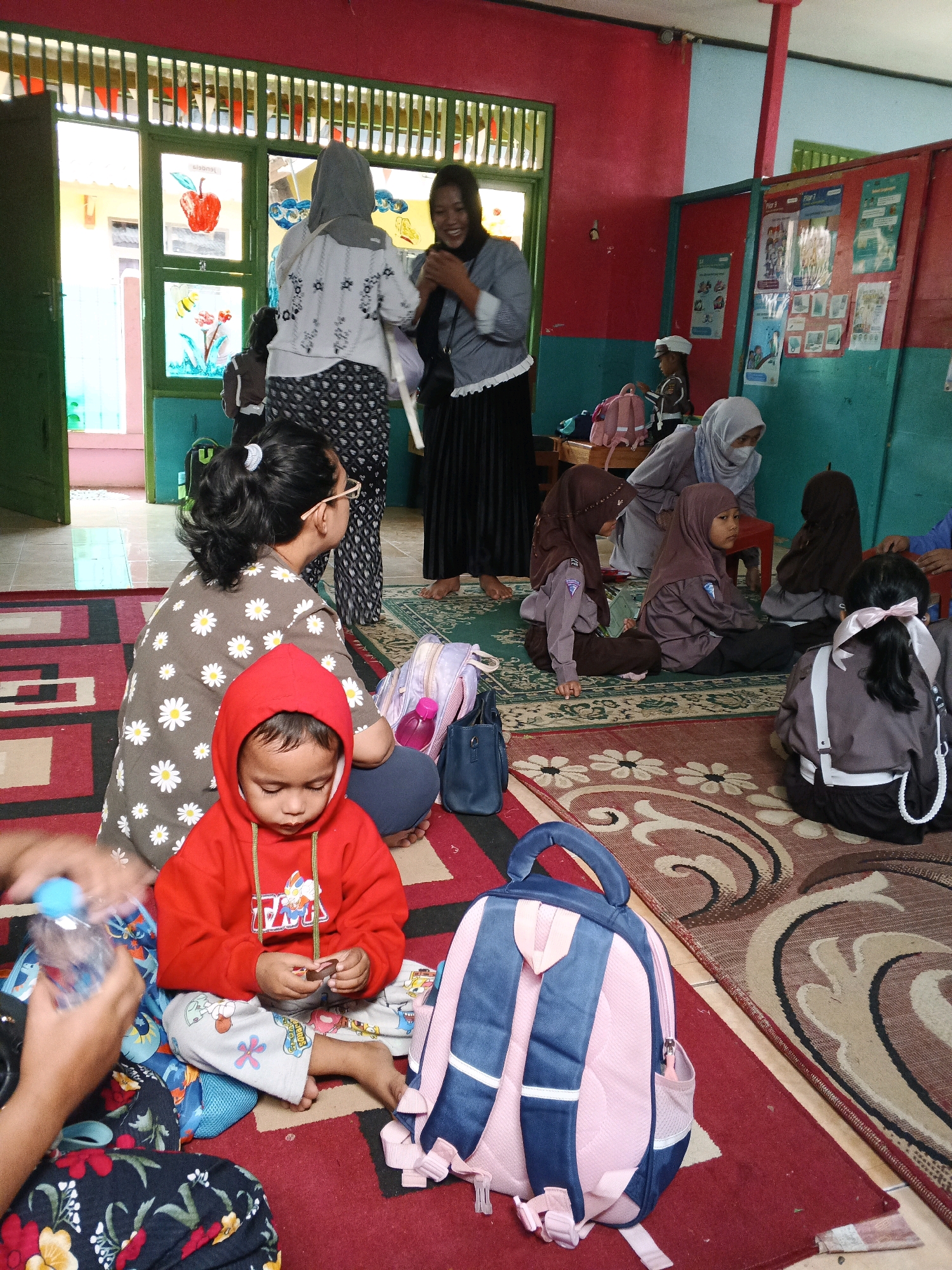 In a brightly lit room with red and teal walls, several people are gathered on a patterned rug. A young child in a red hooded jacket sits on the floor, looking down at something in their hands. To the left, a woman wearing glasses and a floral shirt is seated, also on the floor, with her back to the camera. Other children, some in school uniforms with hijabs, are seated on the rug in small groups, facing towards the back of the room. Two adult women are standing near a window with green trim, one of whom is smiling and speaking. Various backpacks and bags are scattered around the floor. The environment appears to be a classroom or community space, possibly a school in Warungkiara, Indonesia, given the context of the children in uniforms. The overall scene conveys a sense of communal activity and interaction.