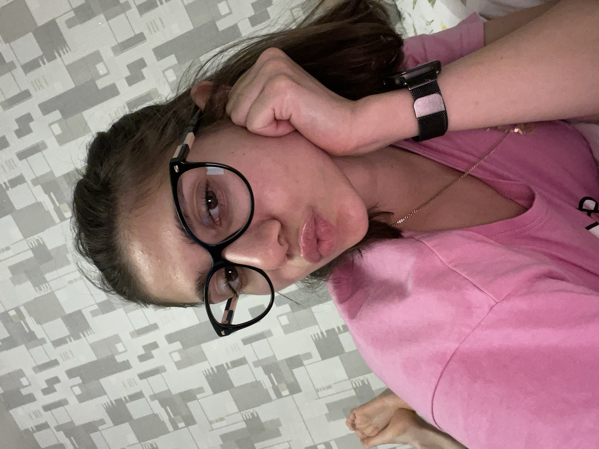 A woman is lying on a bed with a patterned sheet and pillow, wearing a pink t-shirt, black-framed glasses, and a black watch. She rests her face on her fist and purses her lips. There is a gold chain around her neck. The photo appears to have been taken indoors in Sofia, Bulgaria.